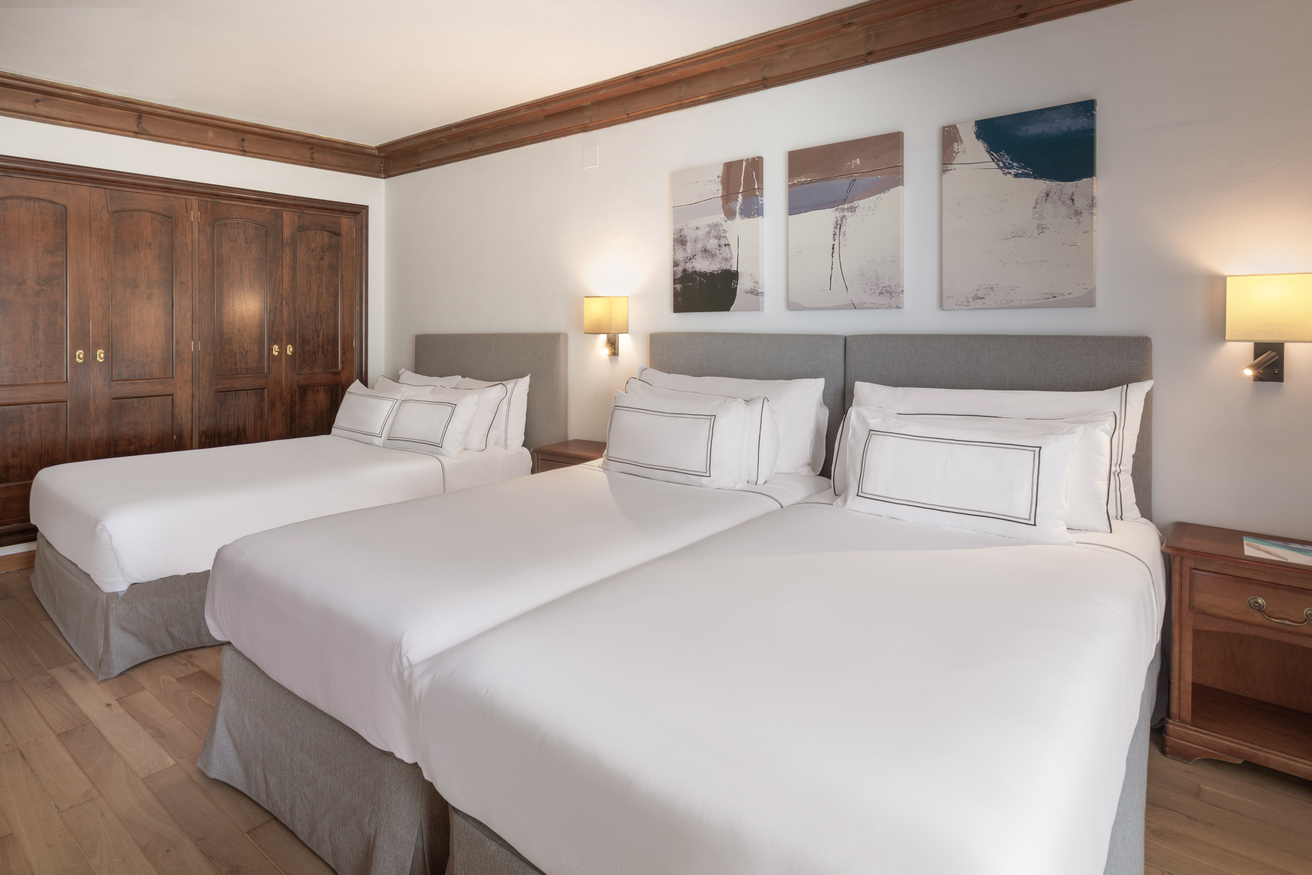 Hotel room with three twin beds, white bedding, wooden closet, and abstract wall art.