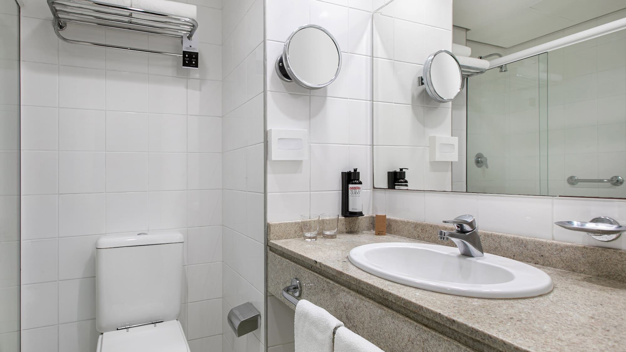 Clean white tiled hotel bathroom with sink, toilet, mirrors, and fresh towels.