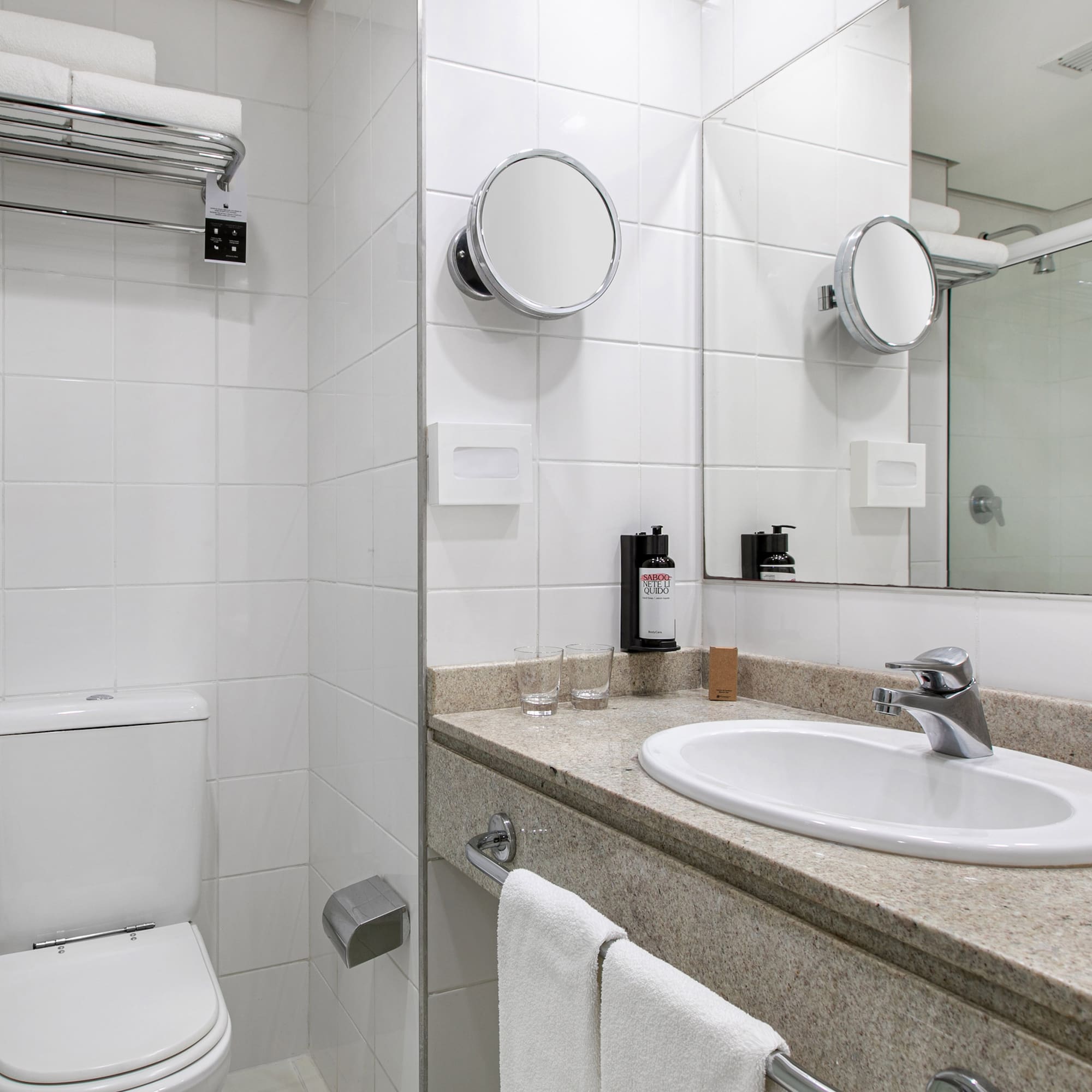 Clean white tiled hotel bathroom with sink, toilet, mirrors, and fresh towels.