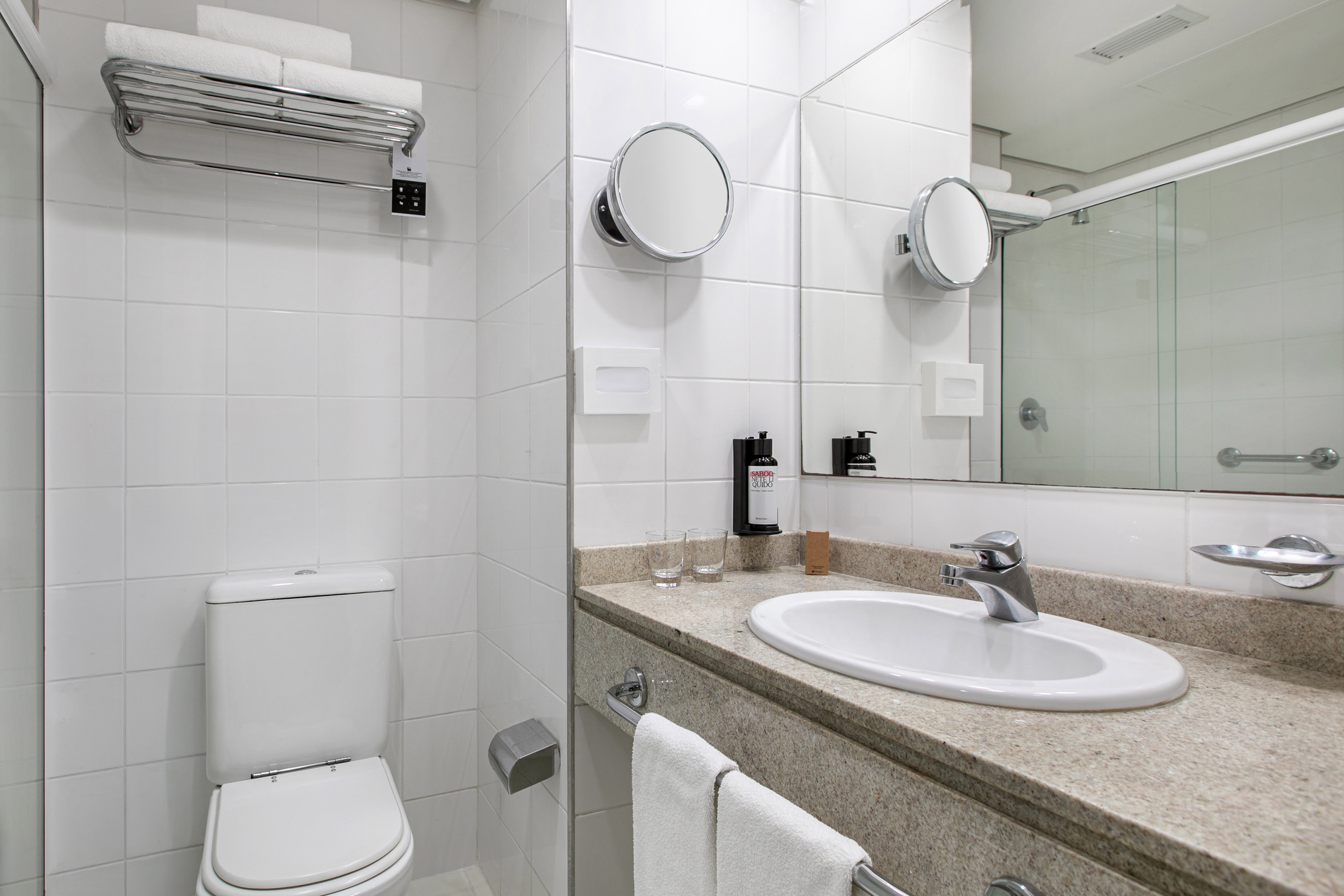 Clean white tiled hotel bathroom with sink, toilet, mirrors, and fresh towels.