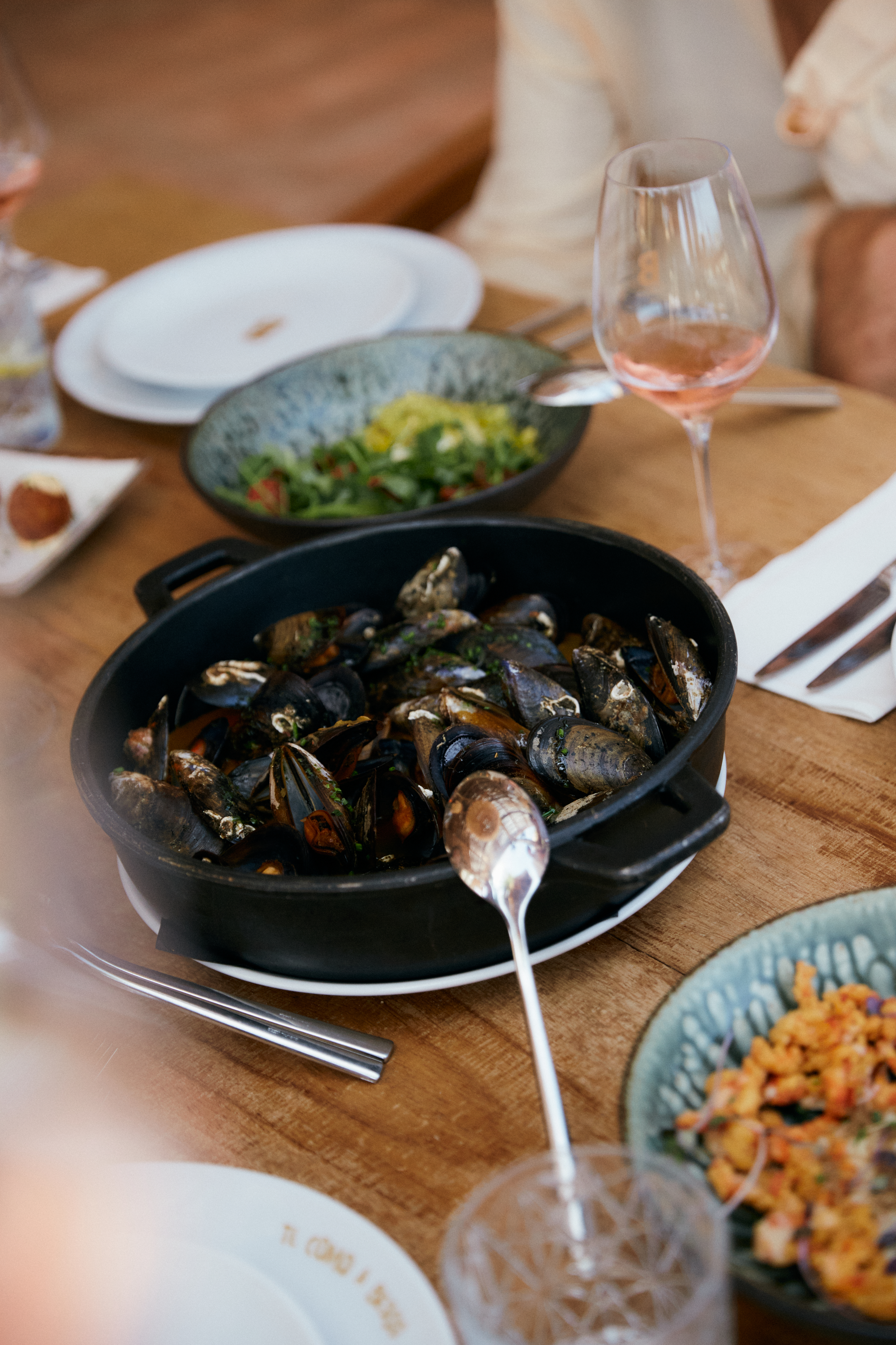 Black pan of steamed mussels, rosé wine, and salad on a wooden table.