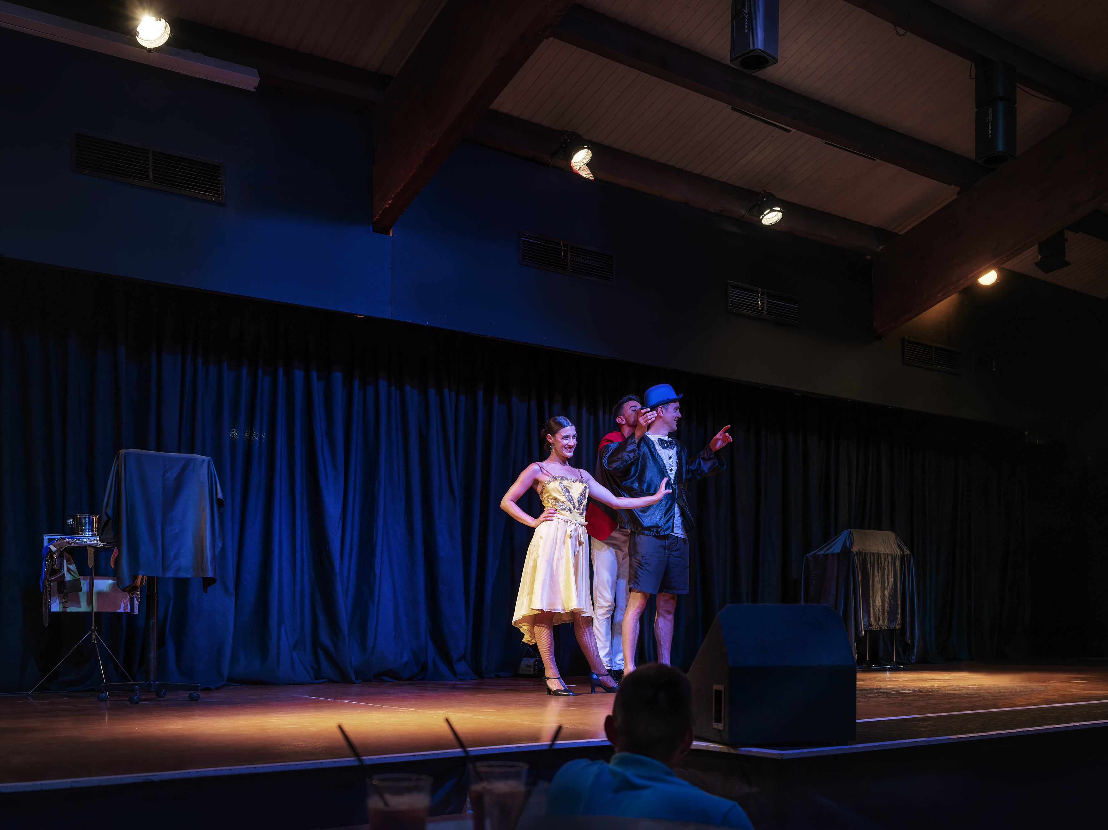 Three entertainers on stage: a woman in a yellow dress and two men, blue curtains.