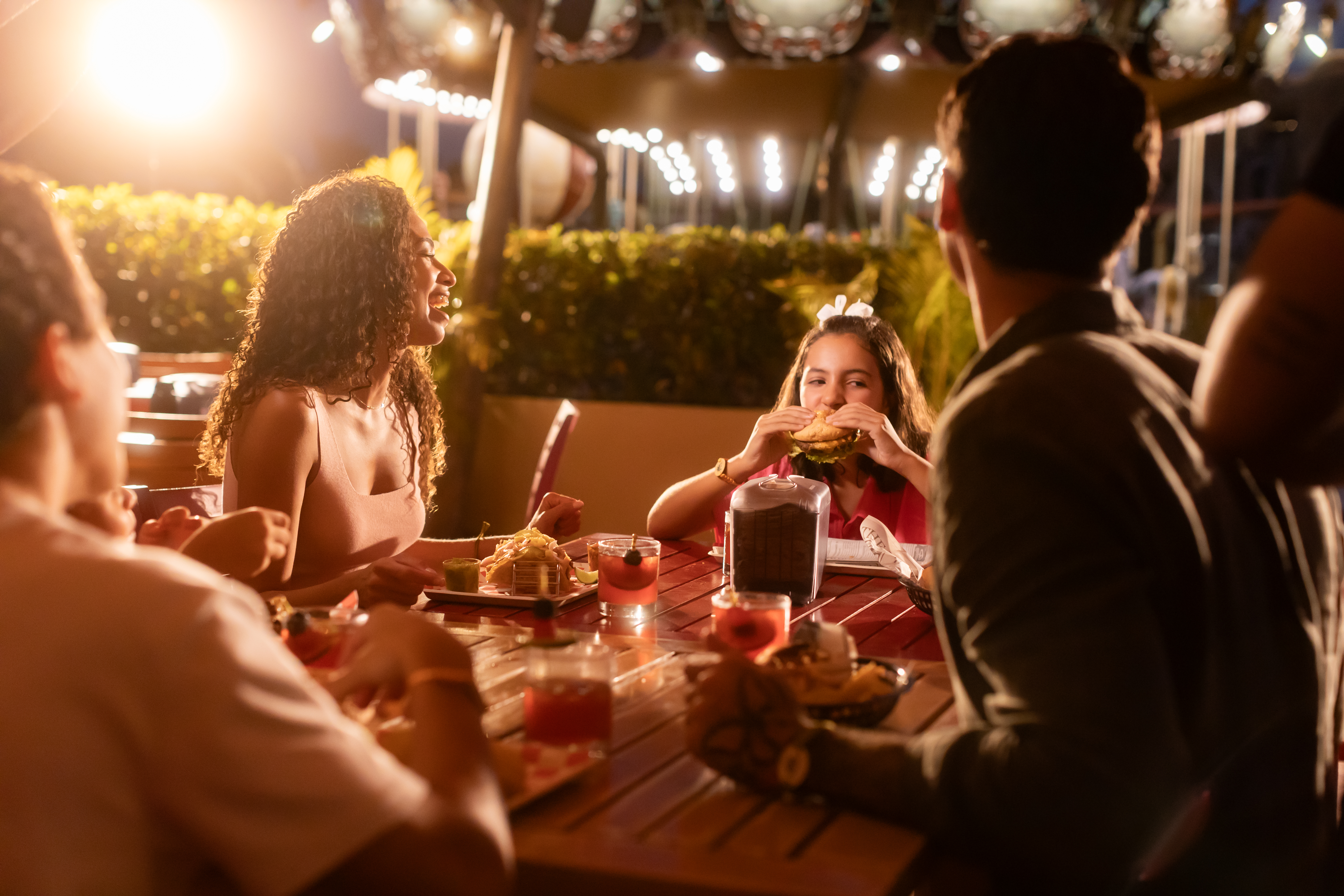 Friends eating burgers & cocktails outdoors at night on an illuminated patio.