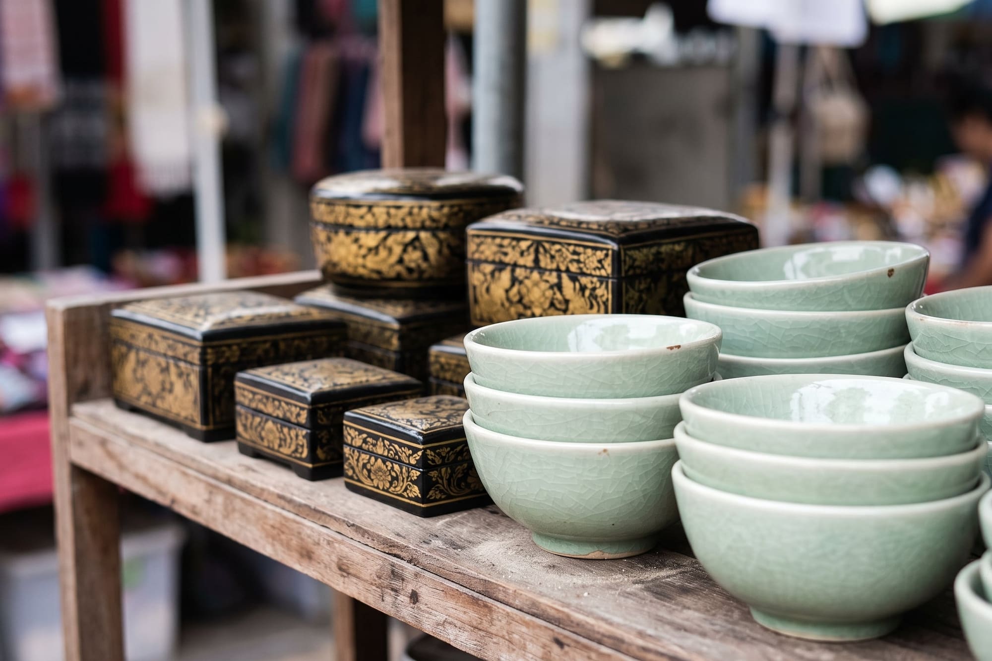 Black and gold lacquered boxes with stacked celadon bowls on a market stall.