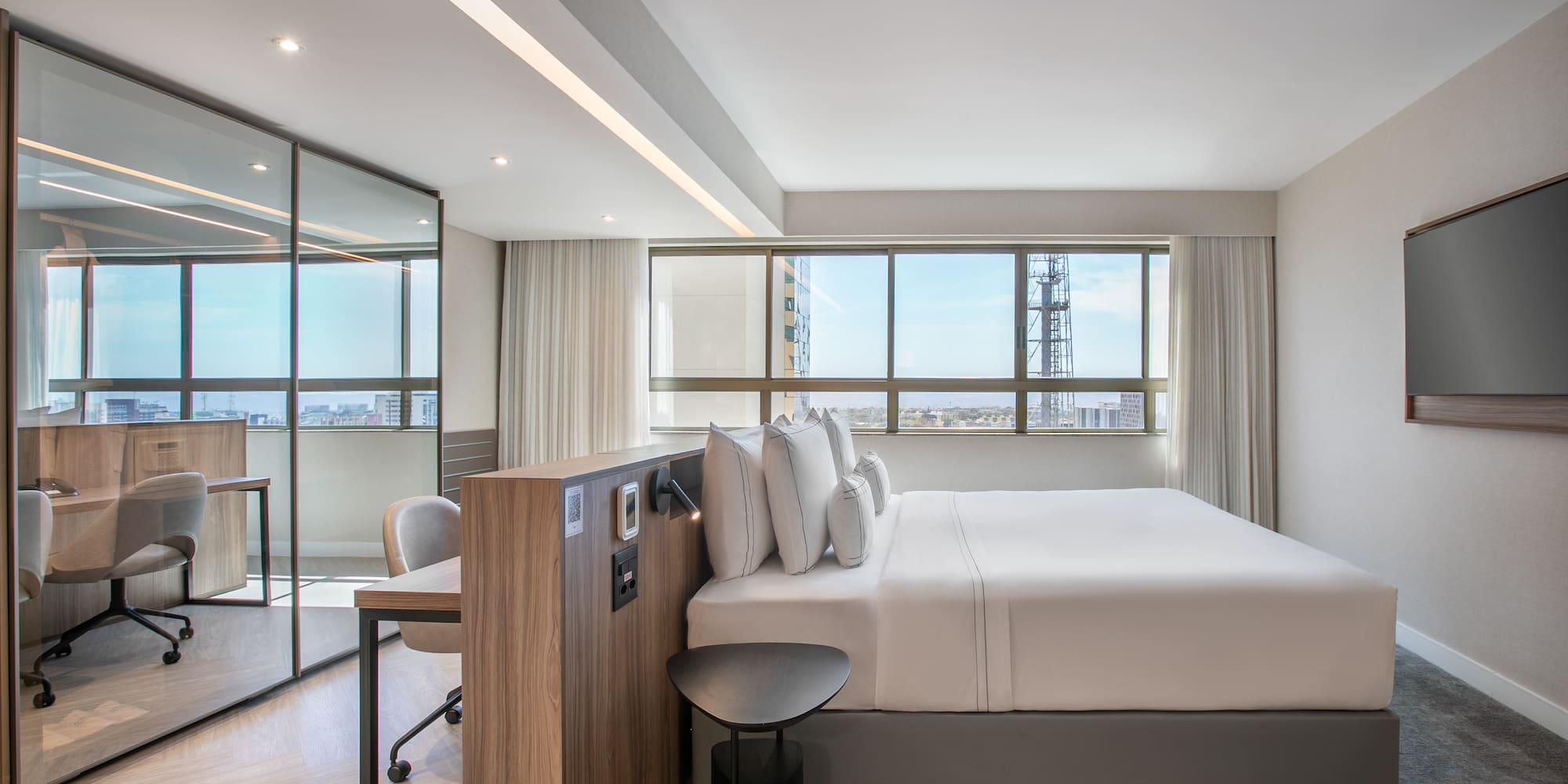 Modern hotel suite with king bed, city view, and glass-partitioned workspace.