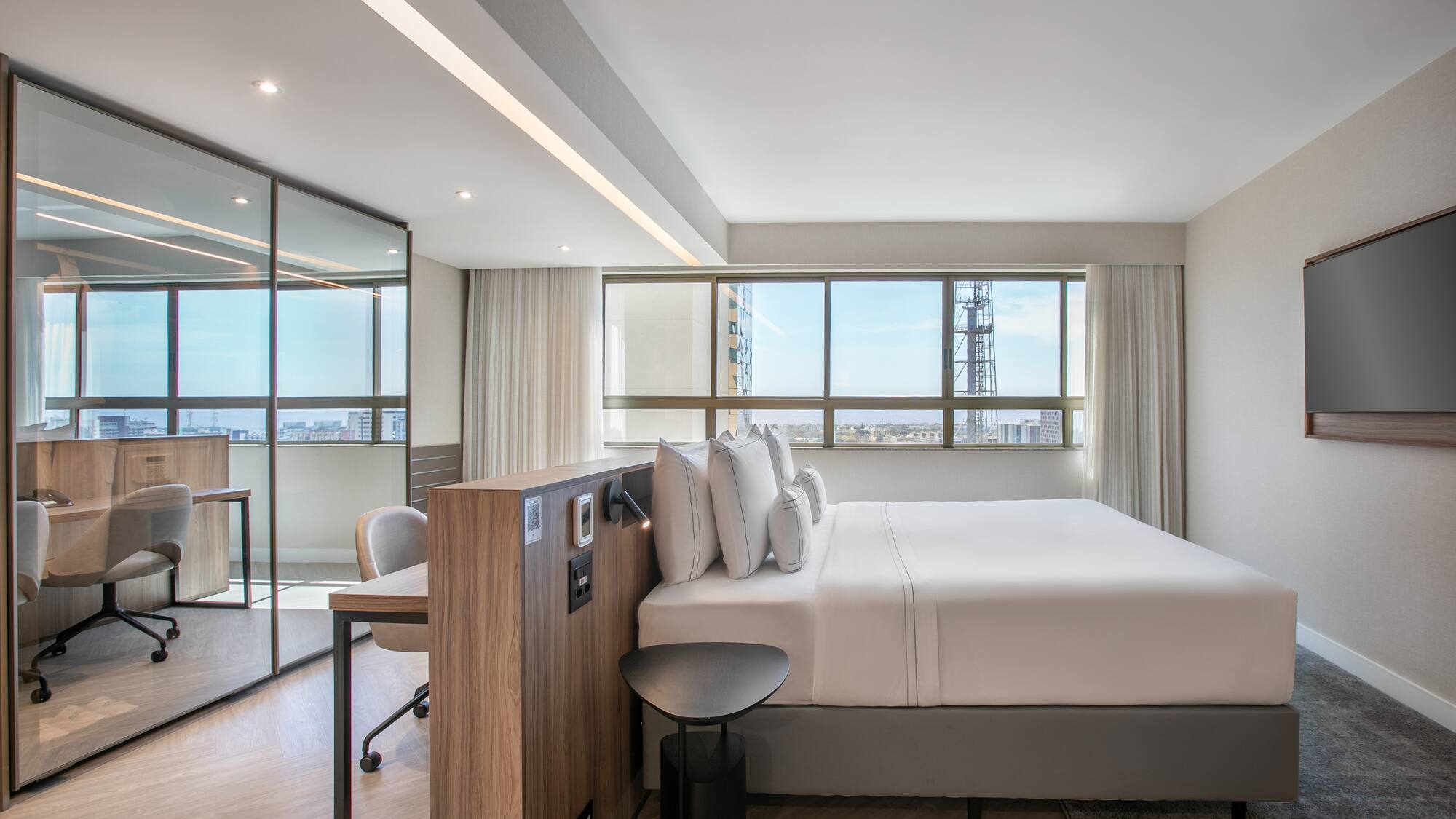Modern hotel suite with king bed, city view, and glass-partitioned workspace.