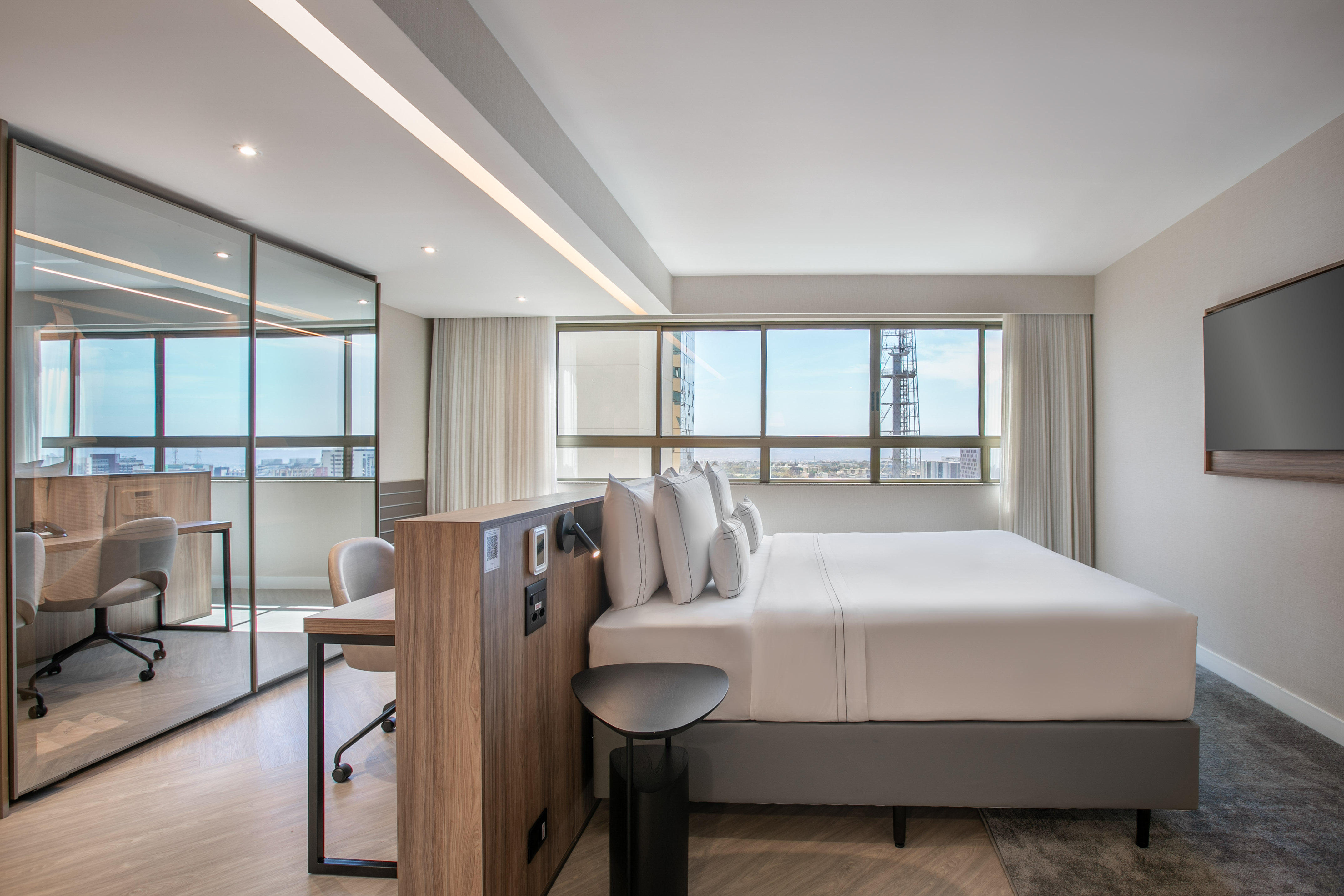 Modern hotel suite with king bed, city view, and glass-partitioned workspace.
