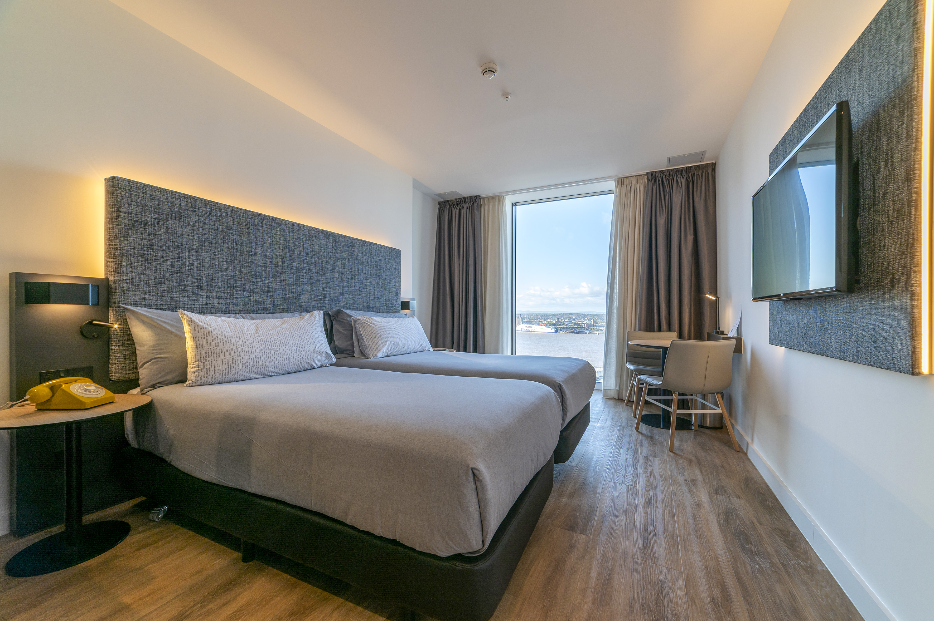 Modern twin hotel room with city waterfront view, TV, and work desk.