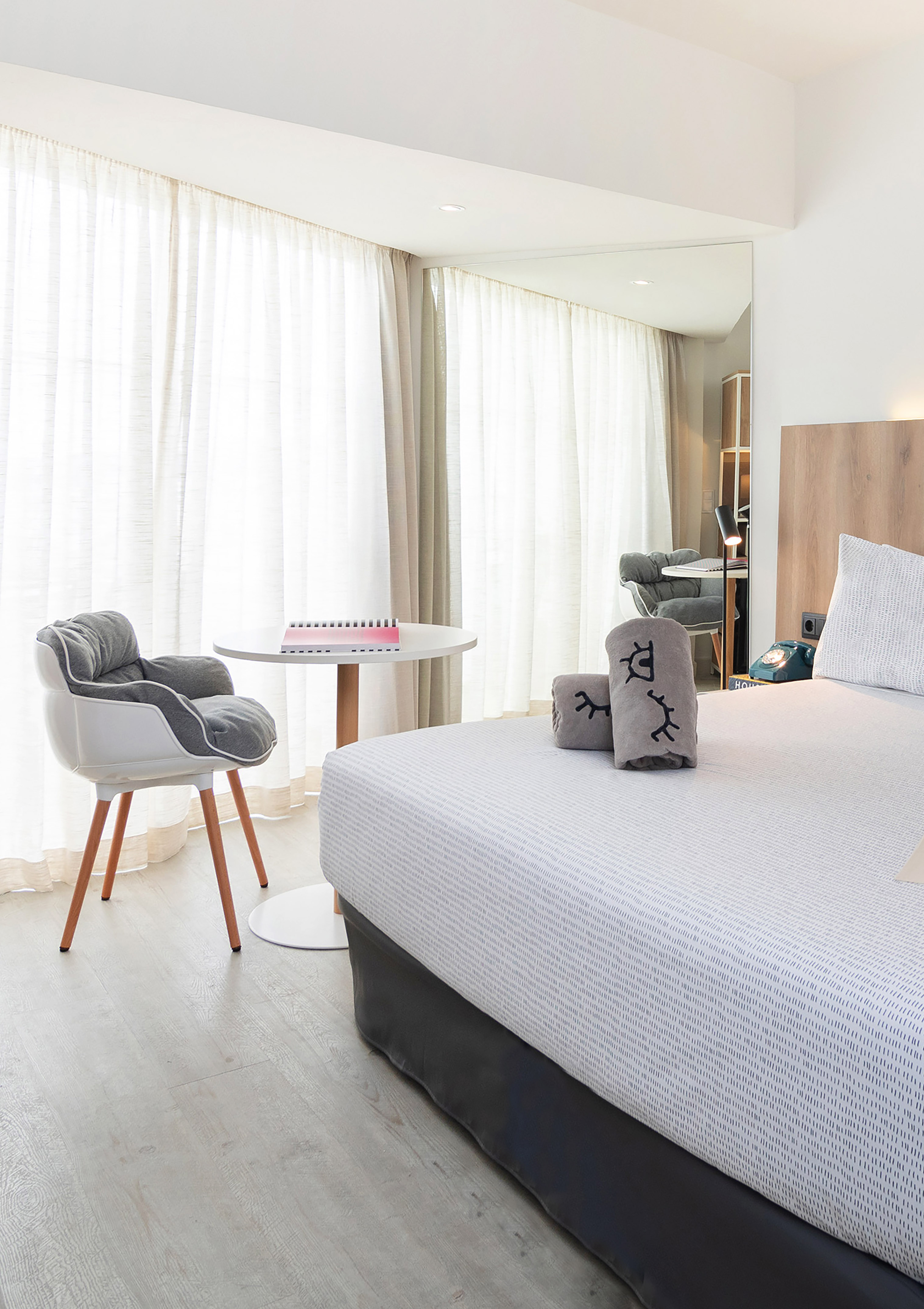 Bright modern hotel room with a bed, grey chair, and sheer window curtains.