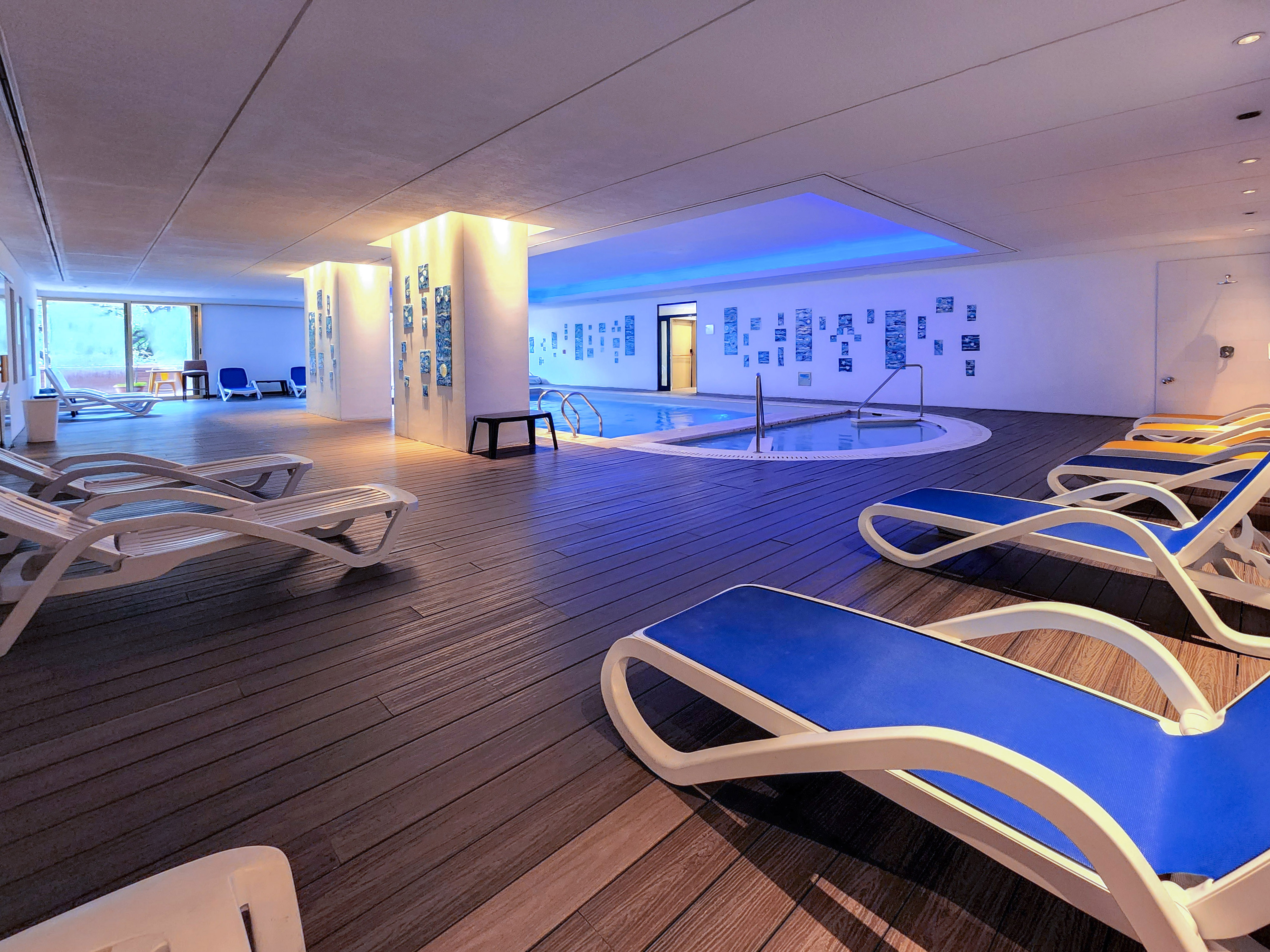 Indoor hotel pool area with blue lighted water, lounge chairs on wooden deck.