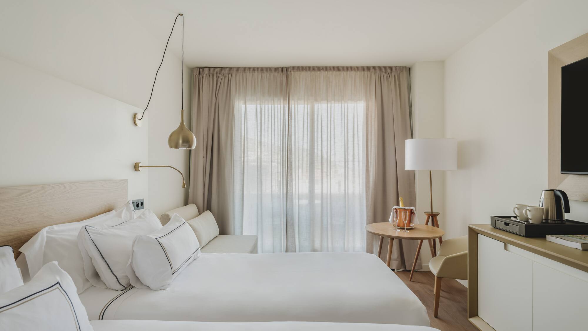 Habitación doble de hotel moderna, tonos neutros, camas y ventanal con cortinas.
