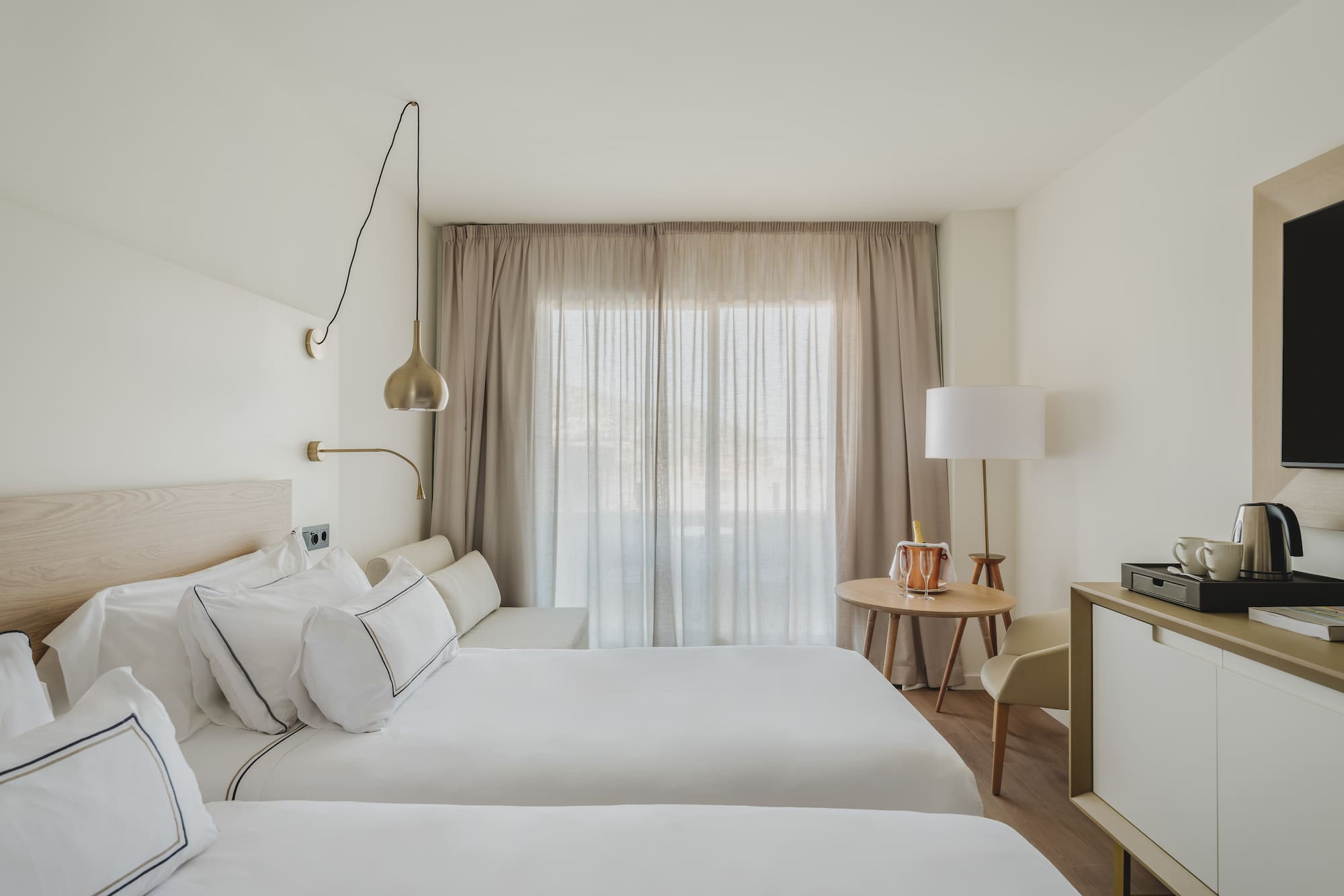 Habitación doble de hotel moderna, tonos neutros, camas y ventanal con cortinas.