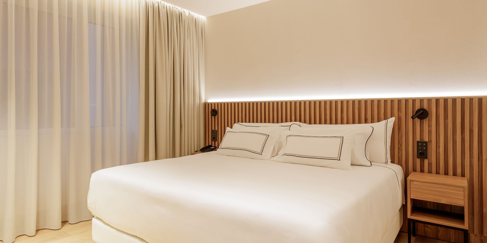 Modern hotel bedroom with large bed, wooden headboard, and soft lighting.