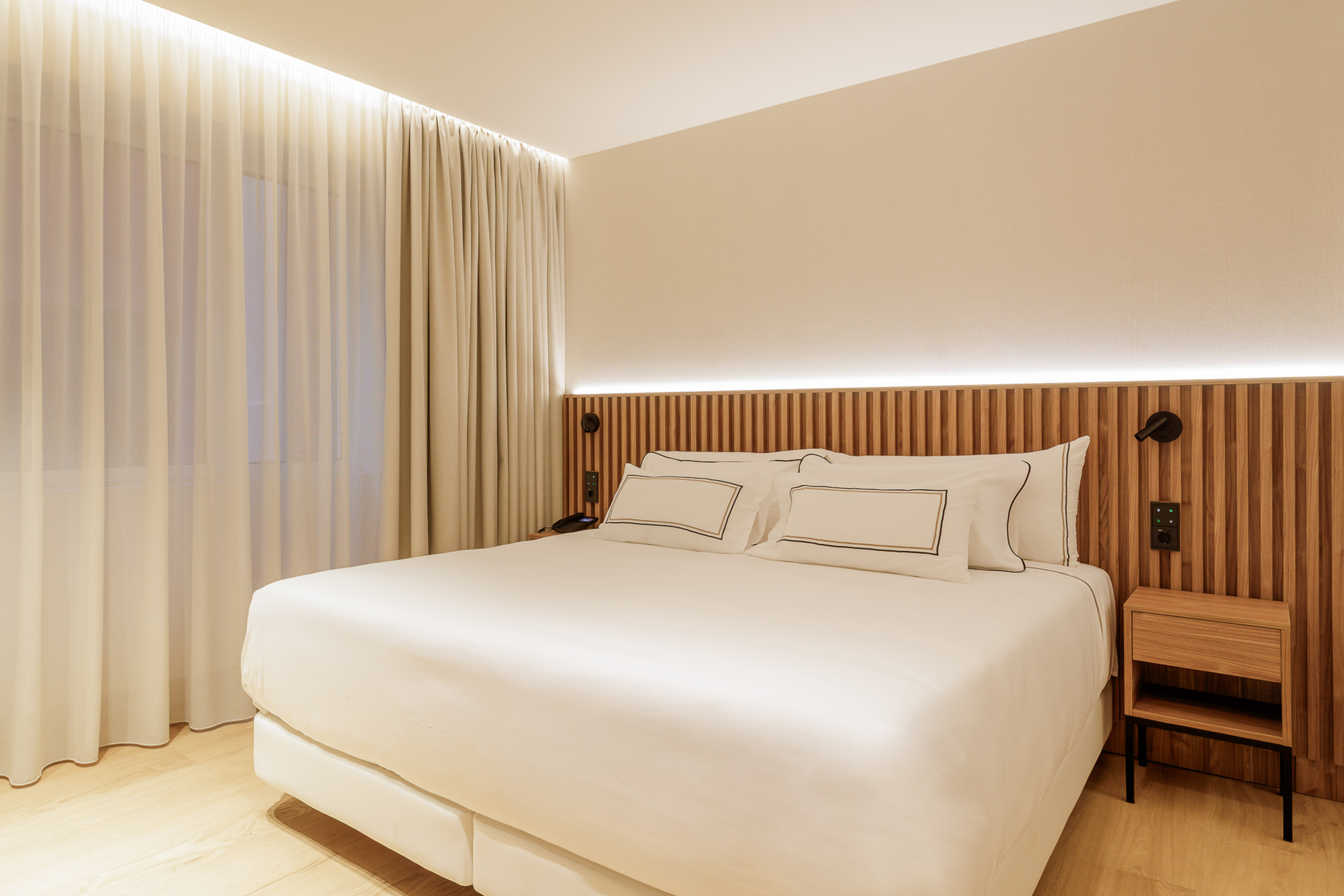 Clean modern hotel bedroom with white bed, wooden headboard, ambient lighting.