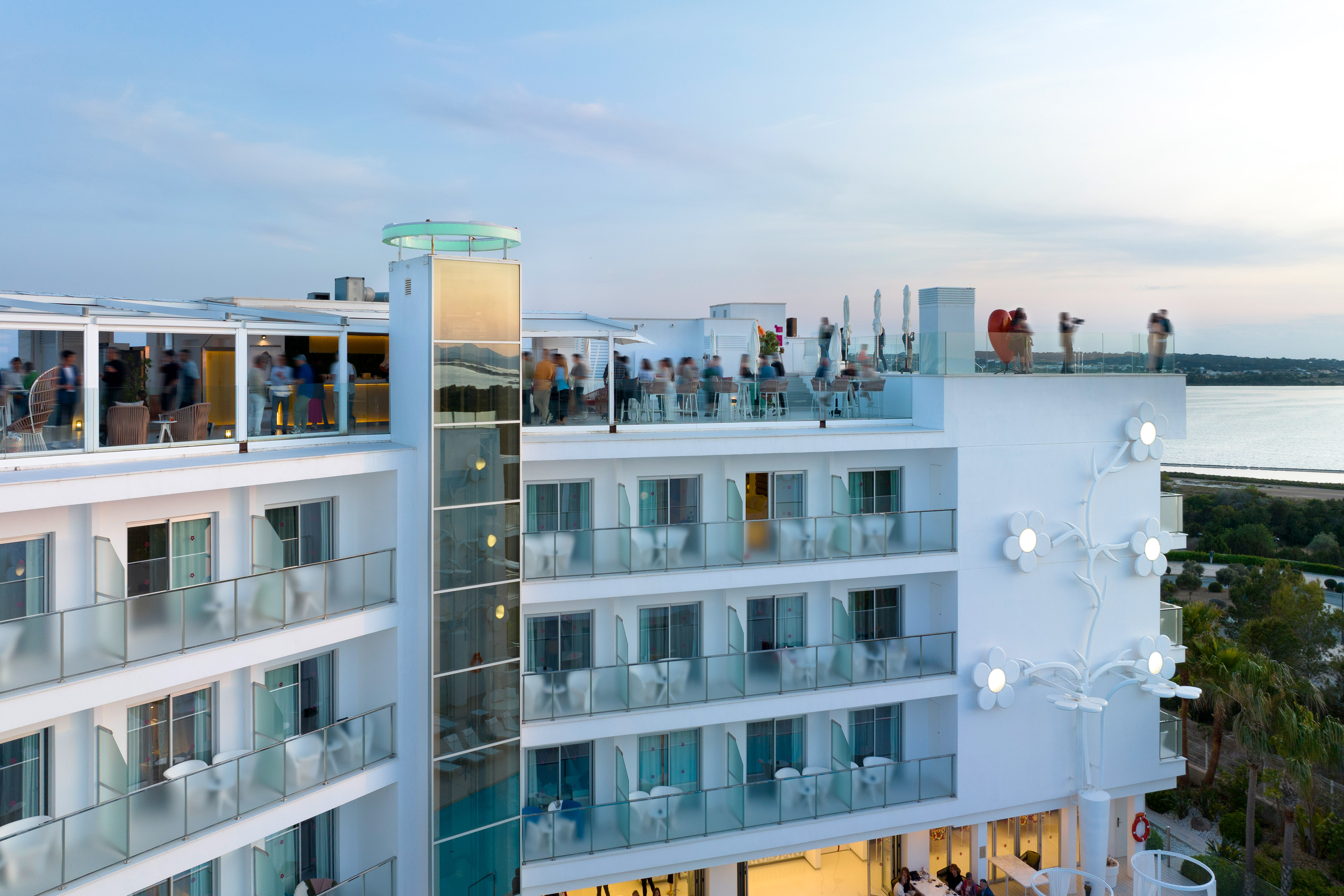Modern white hotel with rooftop bar, glass balconies, floral decor, and sea view.