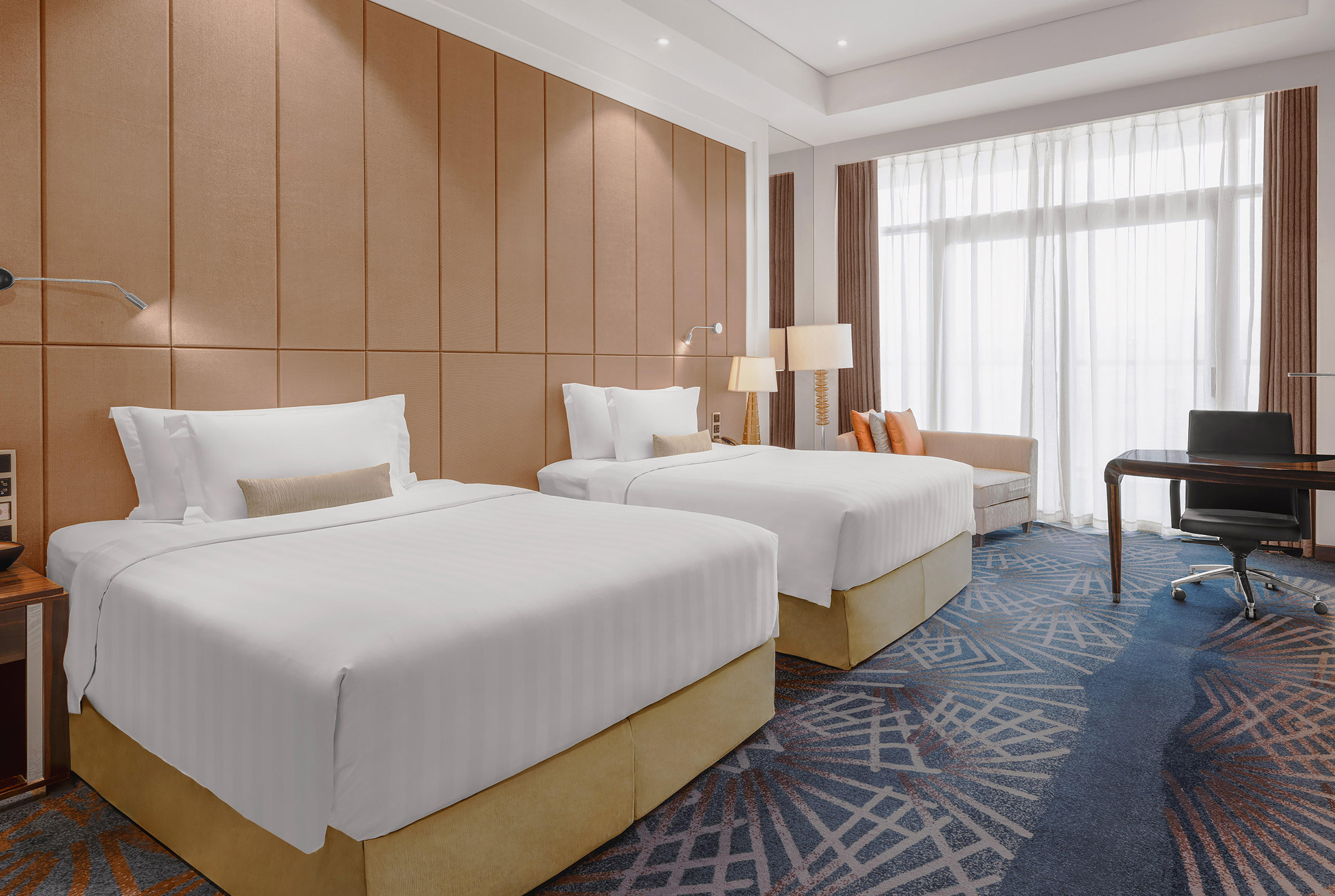 Modern hotel room with two twin beds, blue patterned carpet, desk, and large window.