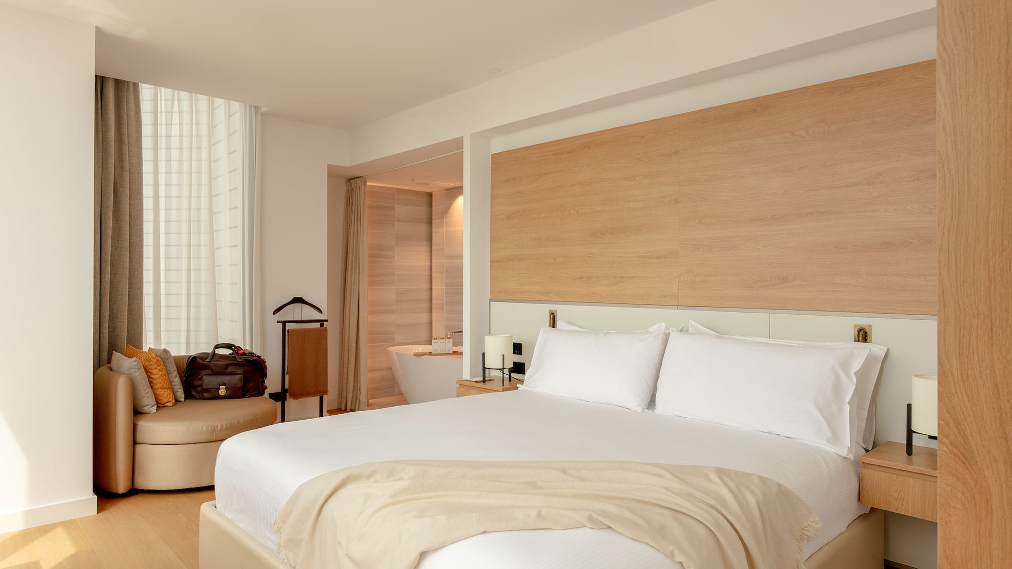 Modern hotel room with white bed, wooden headboard, lounge chair, and bathtub.