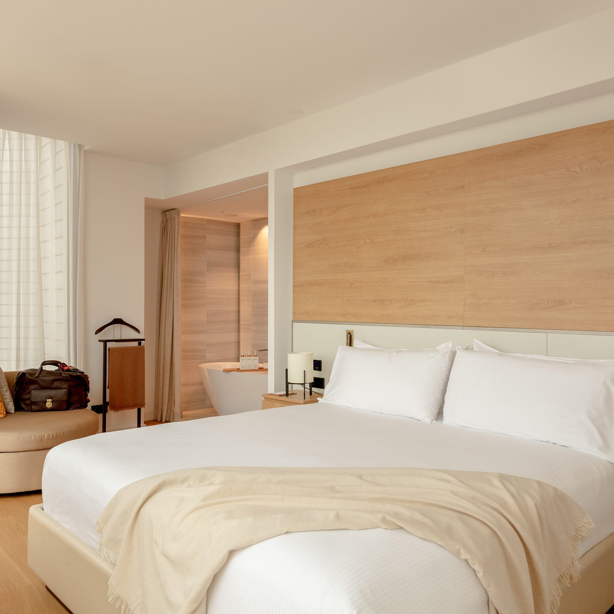 Modern hotel room with white bed, wooden headboard, lounge chair, and bathtub.