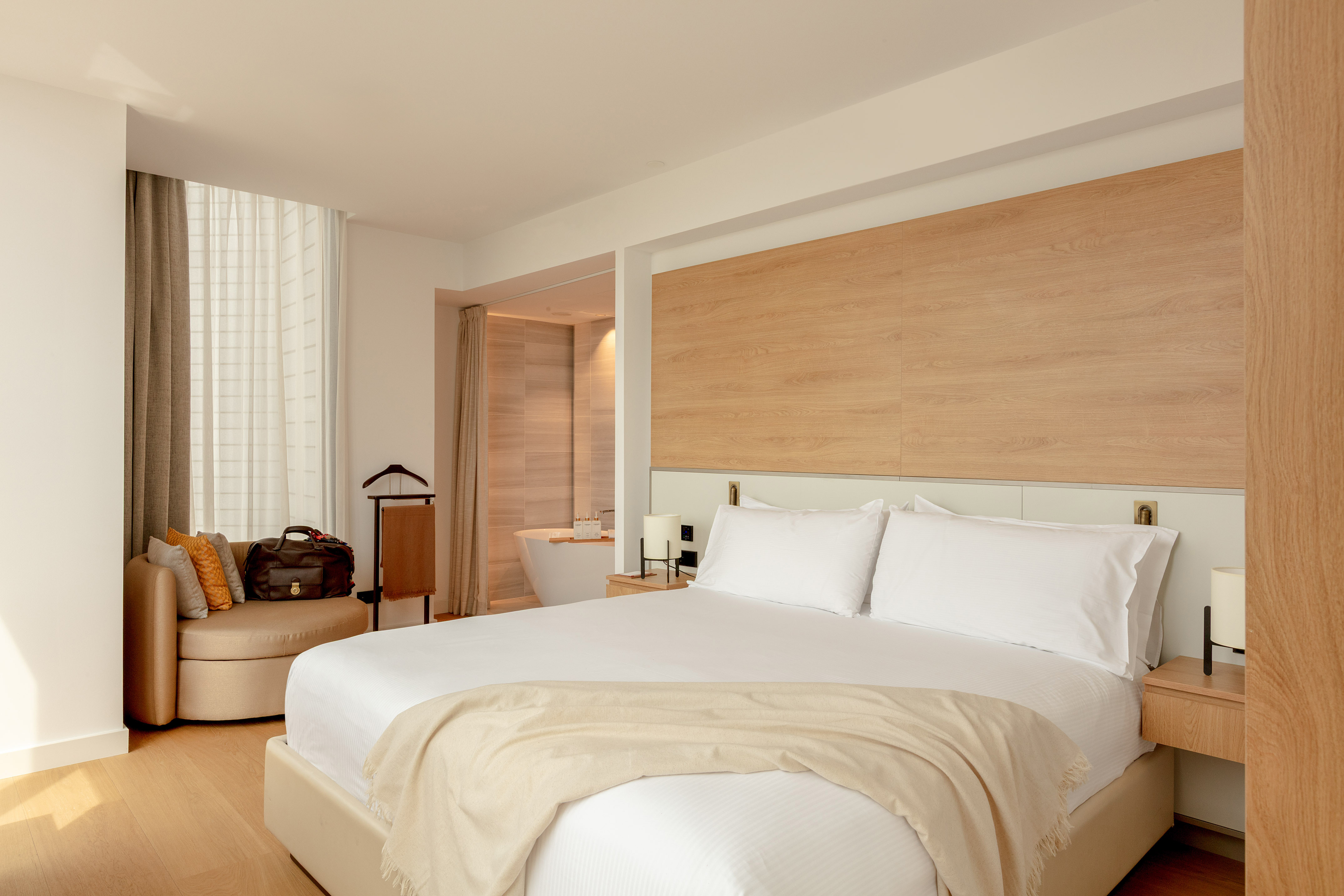 Modern hotel bedroom with white bed, wood accents, and freestanding tub.