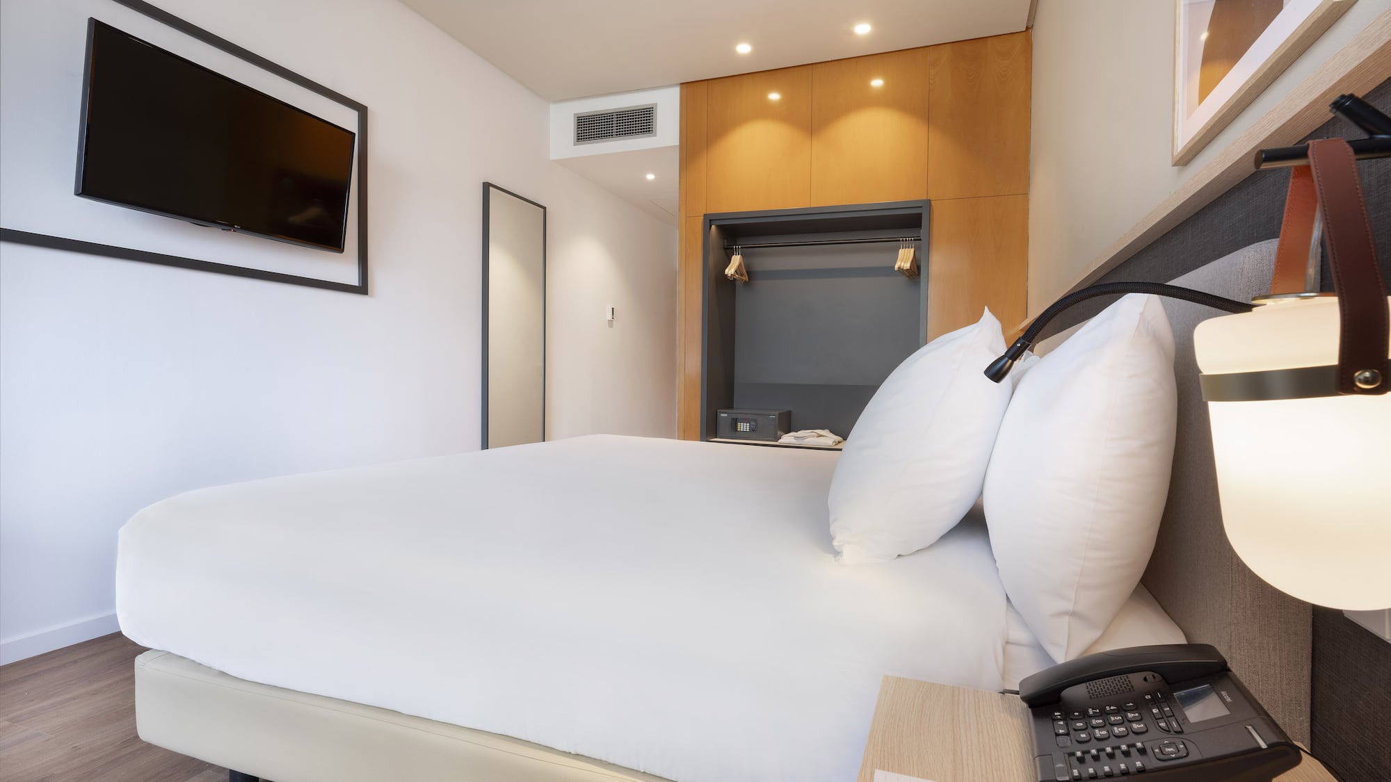 Modern hotel bedroom with white bed, wall-mounted TV, and wooden wardrobe.