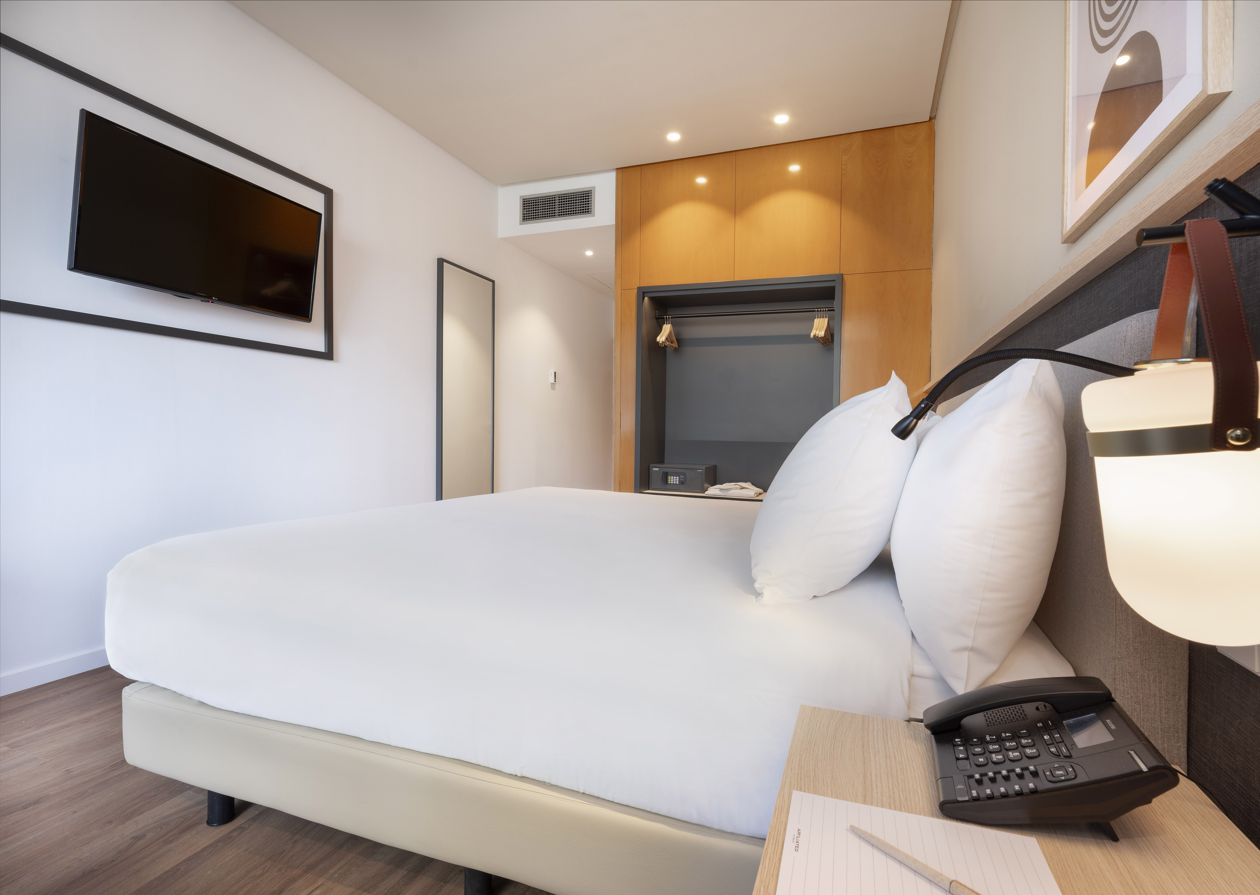 Modern hotel bedroom with white bed, wall-mounted TV, and wooden wardrobe.