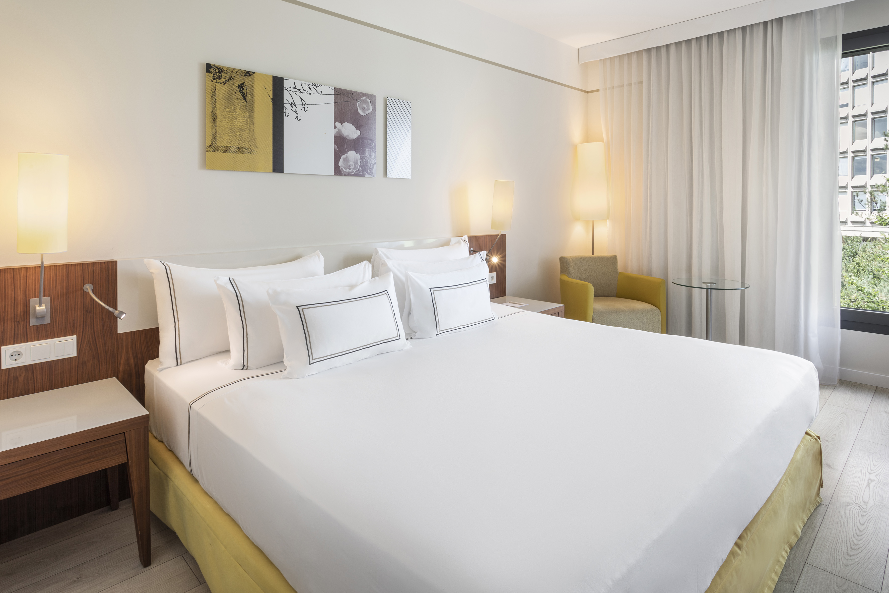 Modern hotel bedroom with white king bed, wooden headboard, yellow armchair, window.