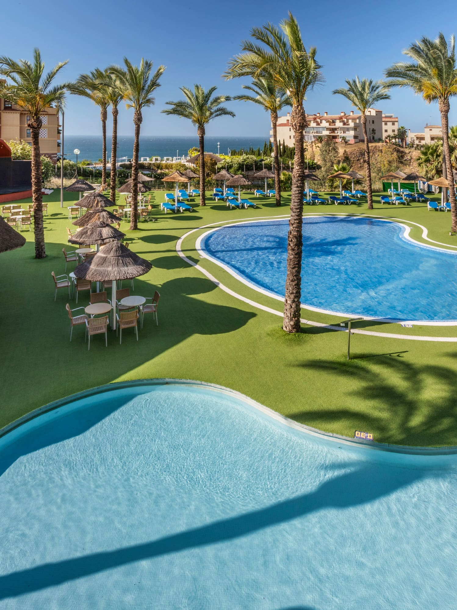 Resort with blue swimming pools, palm trees, thatched bar, and distant ocean.