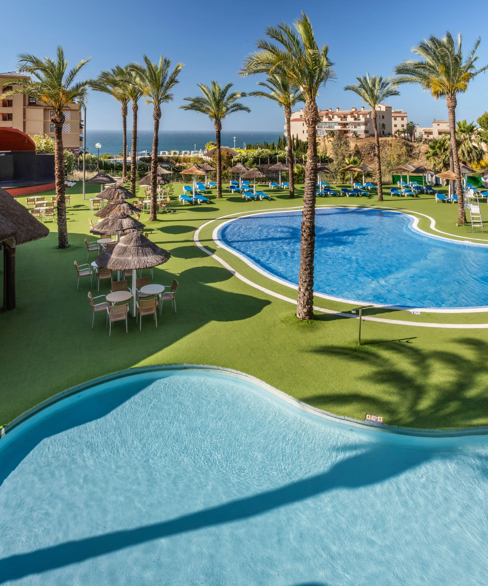 Sunny resort pools, palm trees, poolside bar, hotel, and ocean view.