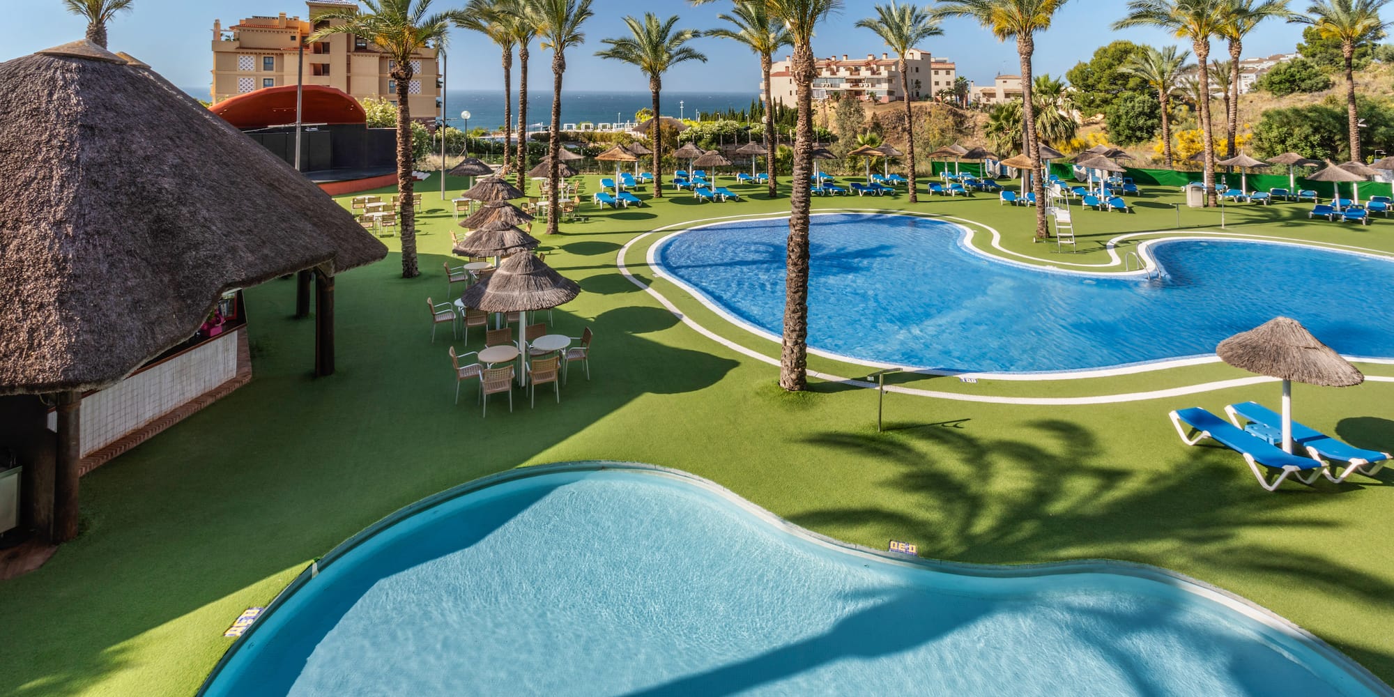Tropical resort pools with palm trees, thatched bar, sun loungers, and ocean.