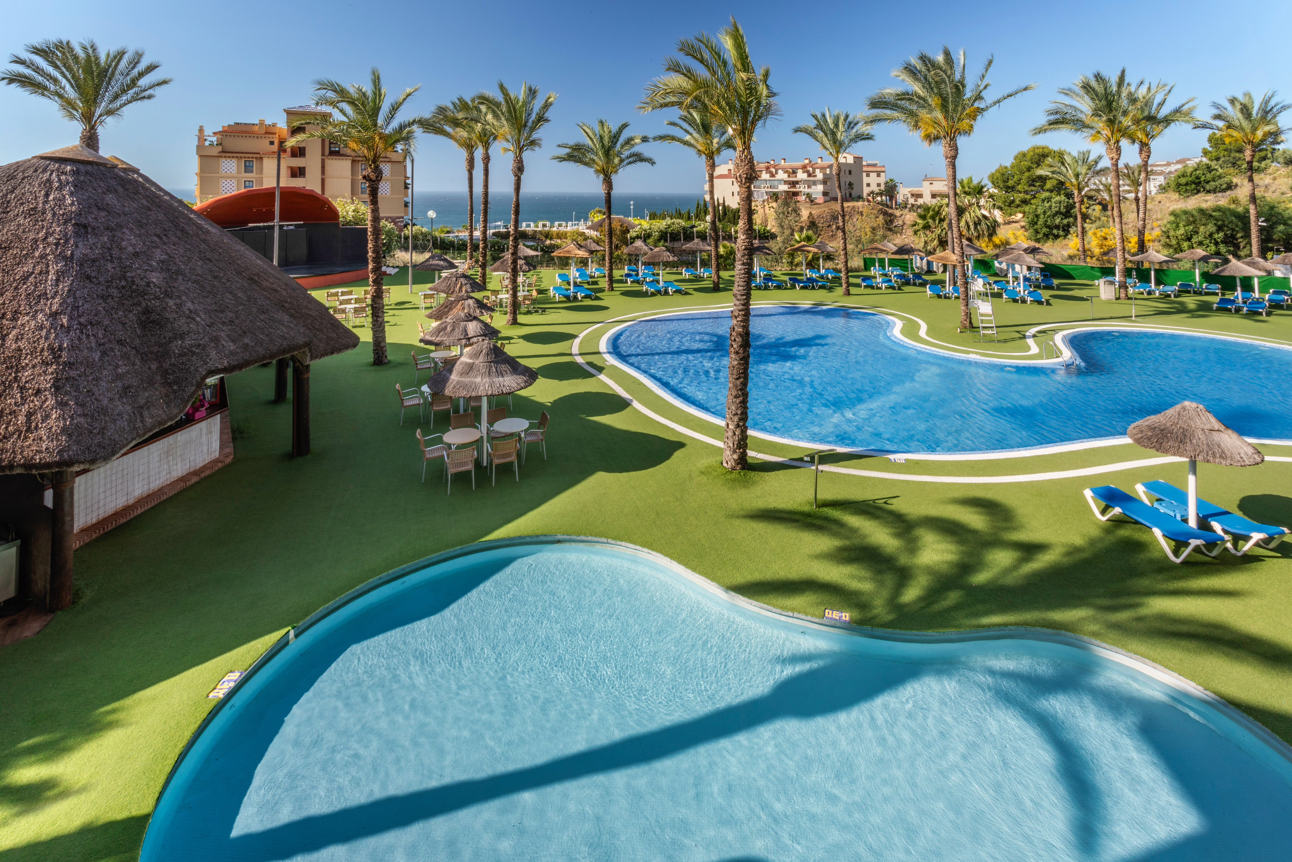 Resort with blue swimming pools, palm trees, thatched bar, and distant ocean.