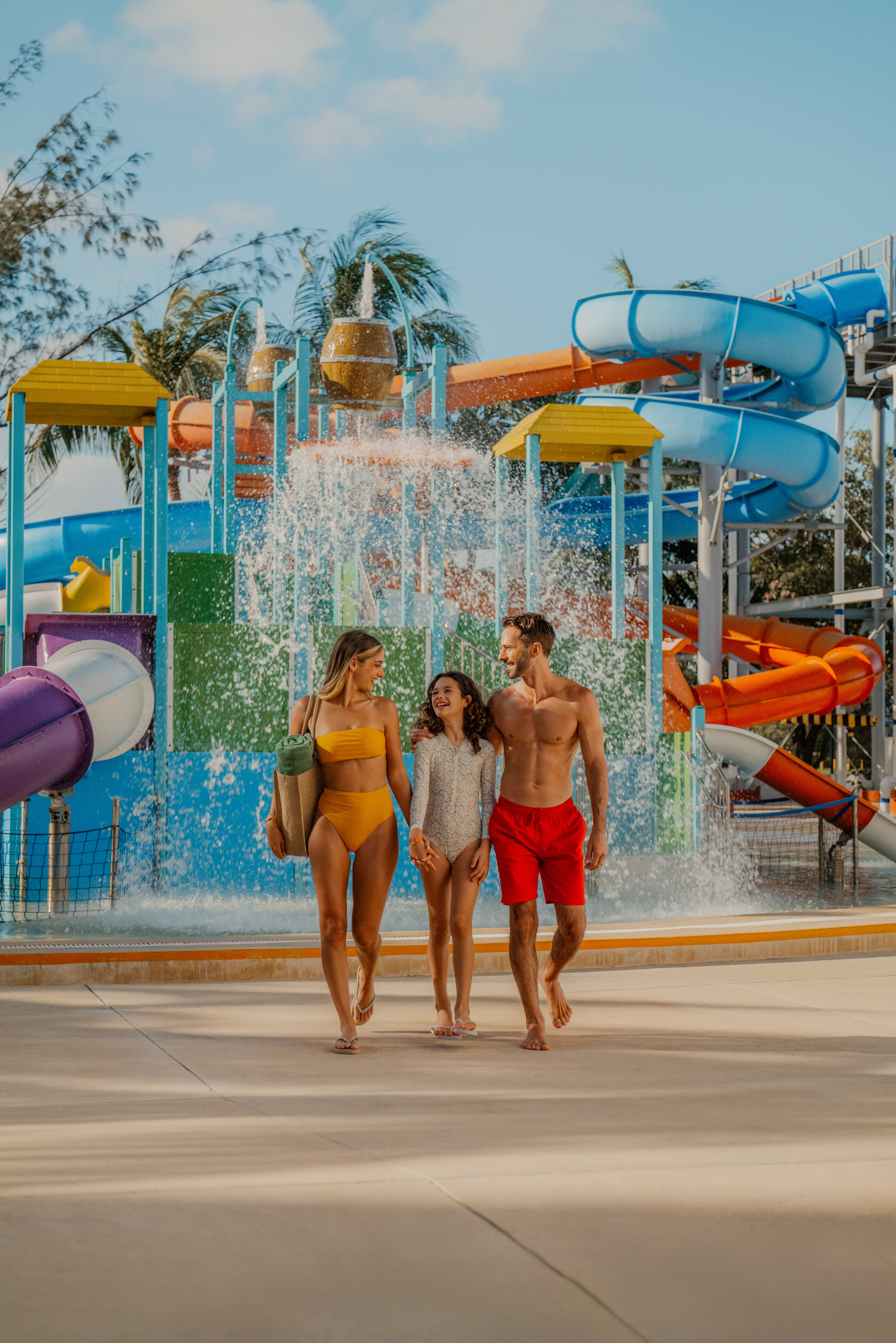 Smiling family in swimsuits walks by colorful water park slides.