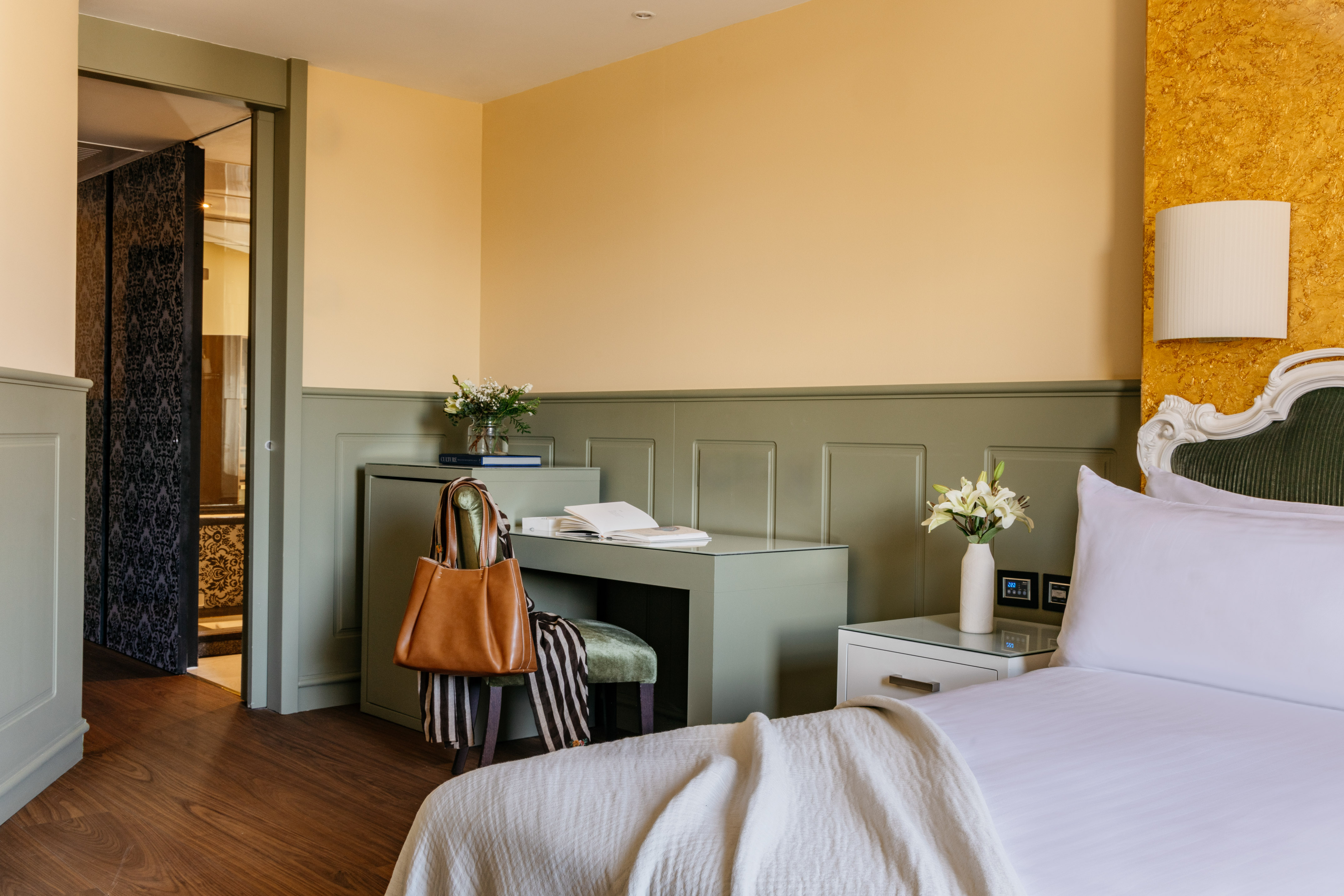 Habitación de hotel elegante con cama, escritorio verde, paredes amarillas y puerta de baño.