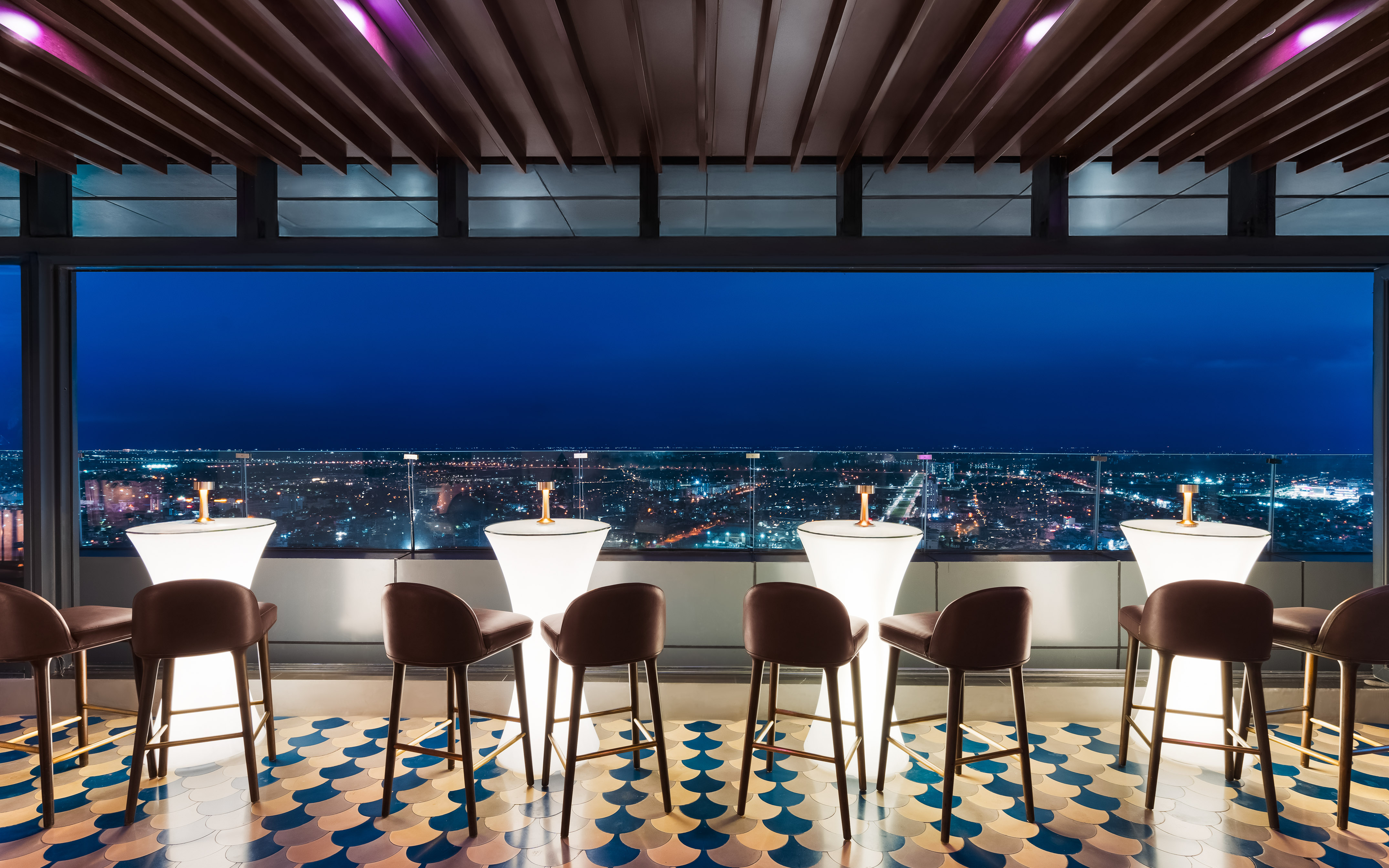 Nighttime rooftop bar with city skyline view, illuminated tables and stools.