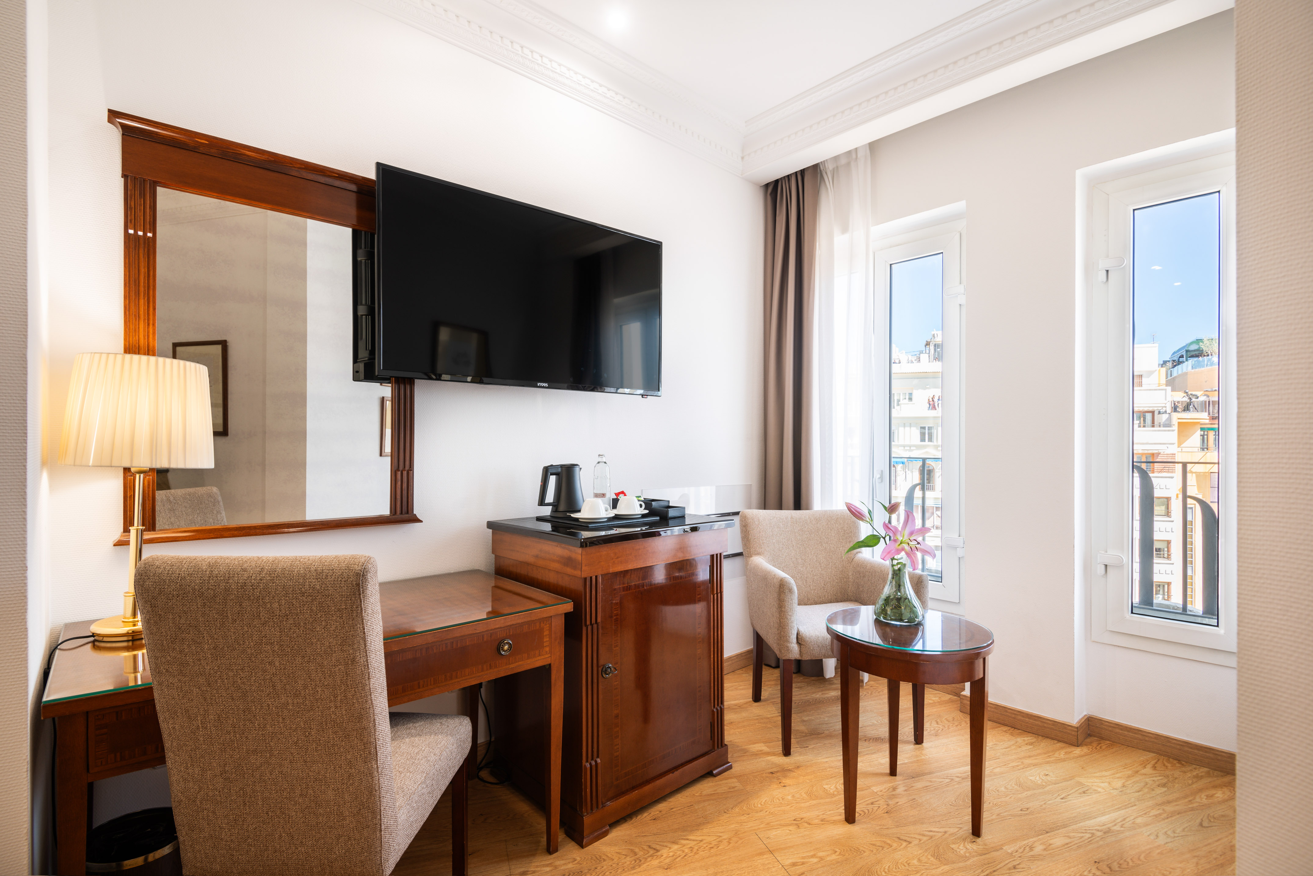 Modern hotel room interior with desk, wall TV, minibar, and bright windows.