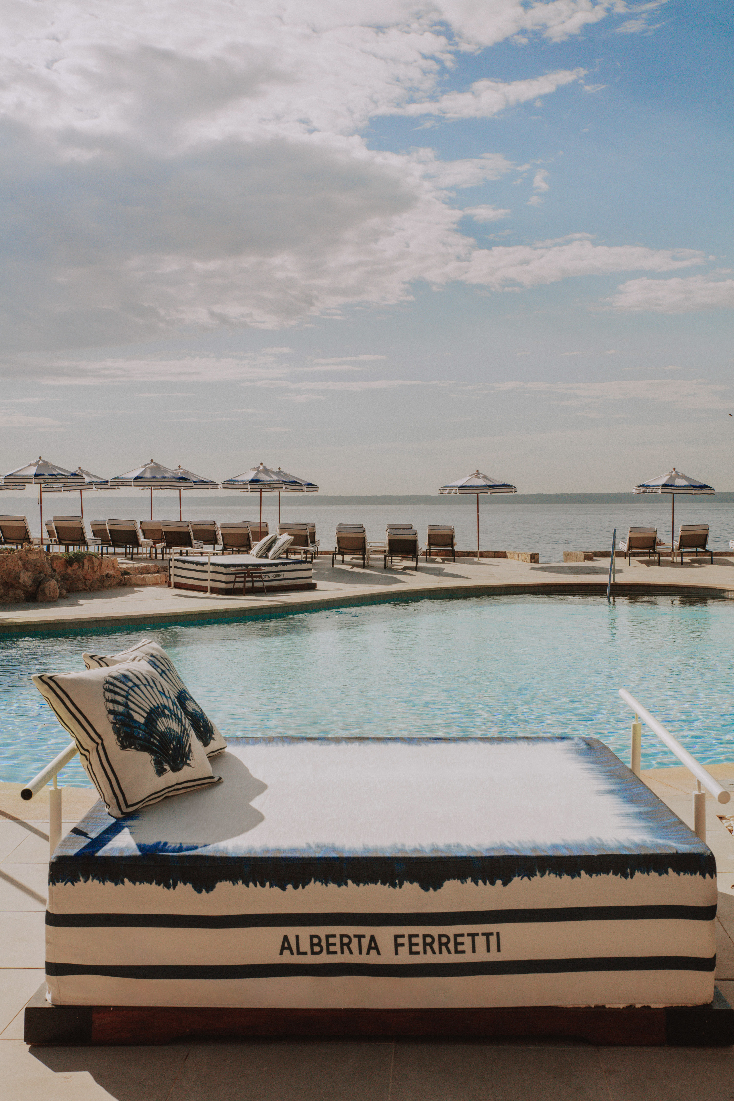 Alberta Ferretti daybed by resort pool, ocean view, striped umbrellas.