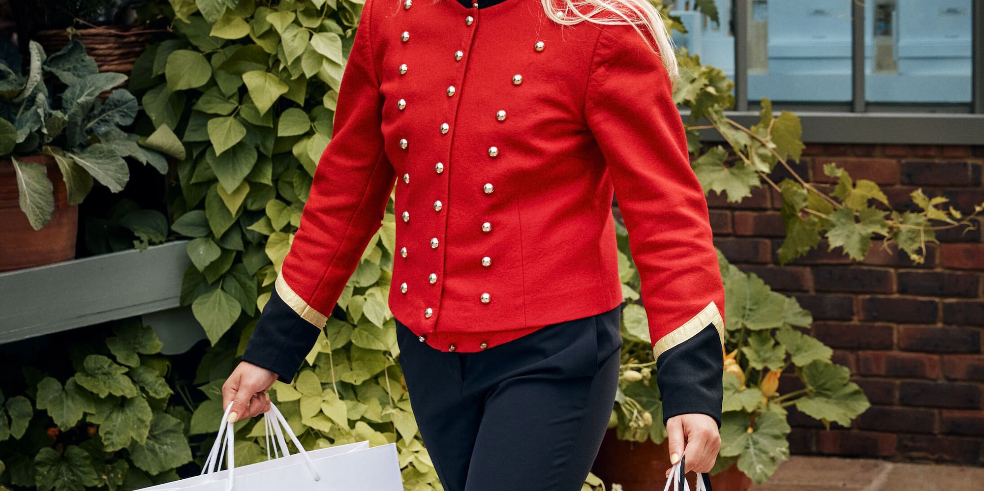 Woman in red Bicester Village uniform carrying Ganni and Sandro shopping bags.
