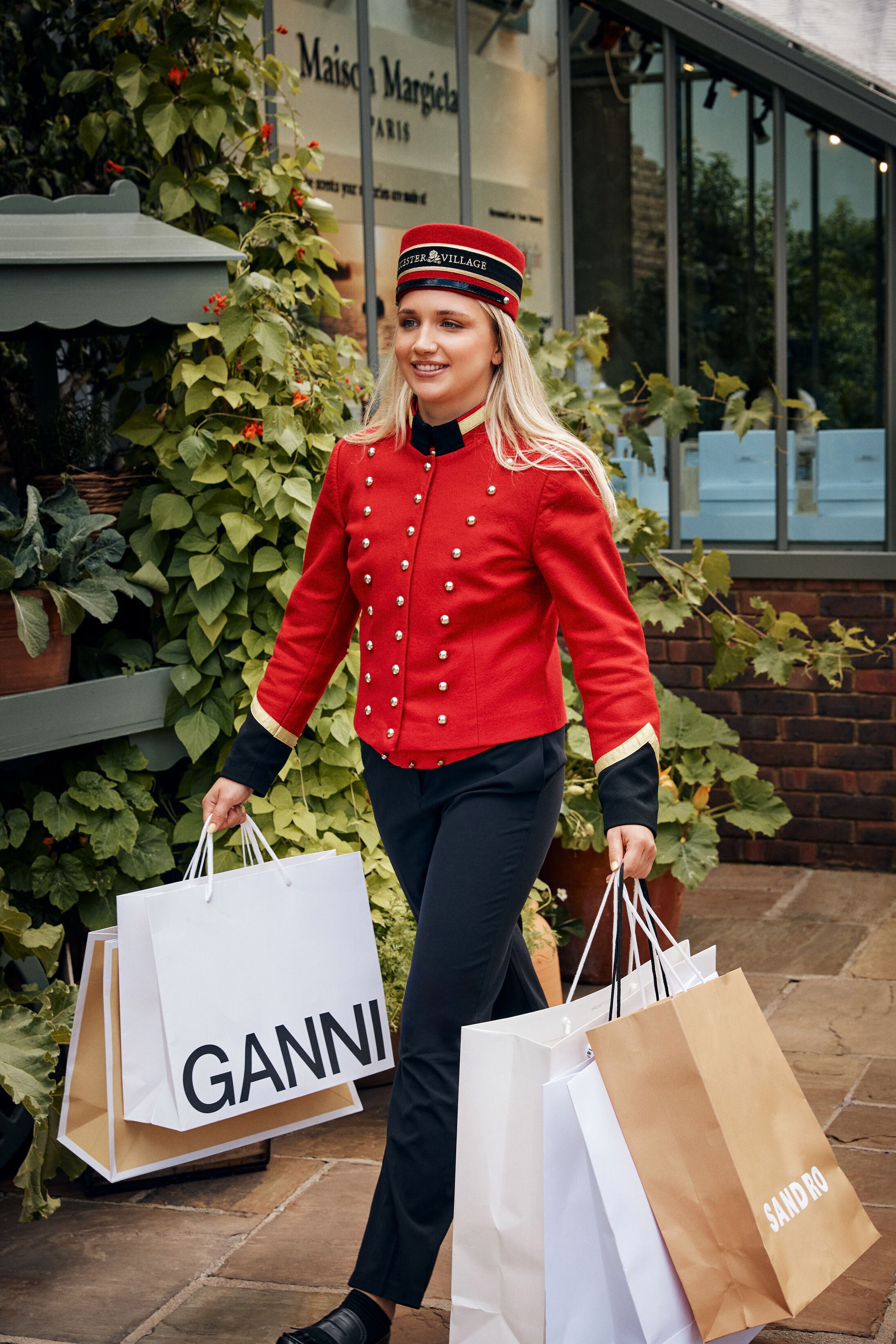 Woman in red Bicester Village uniform carrying Ganni and Sandro shopping bags.