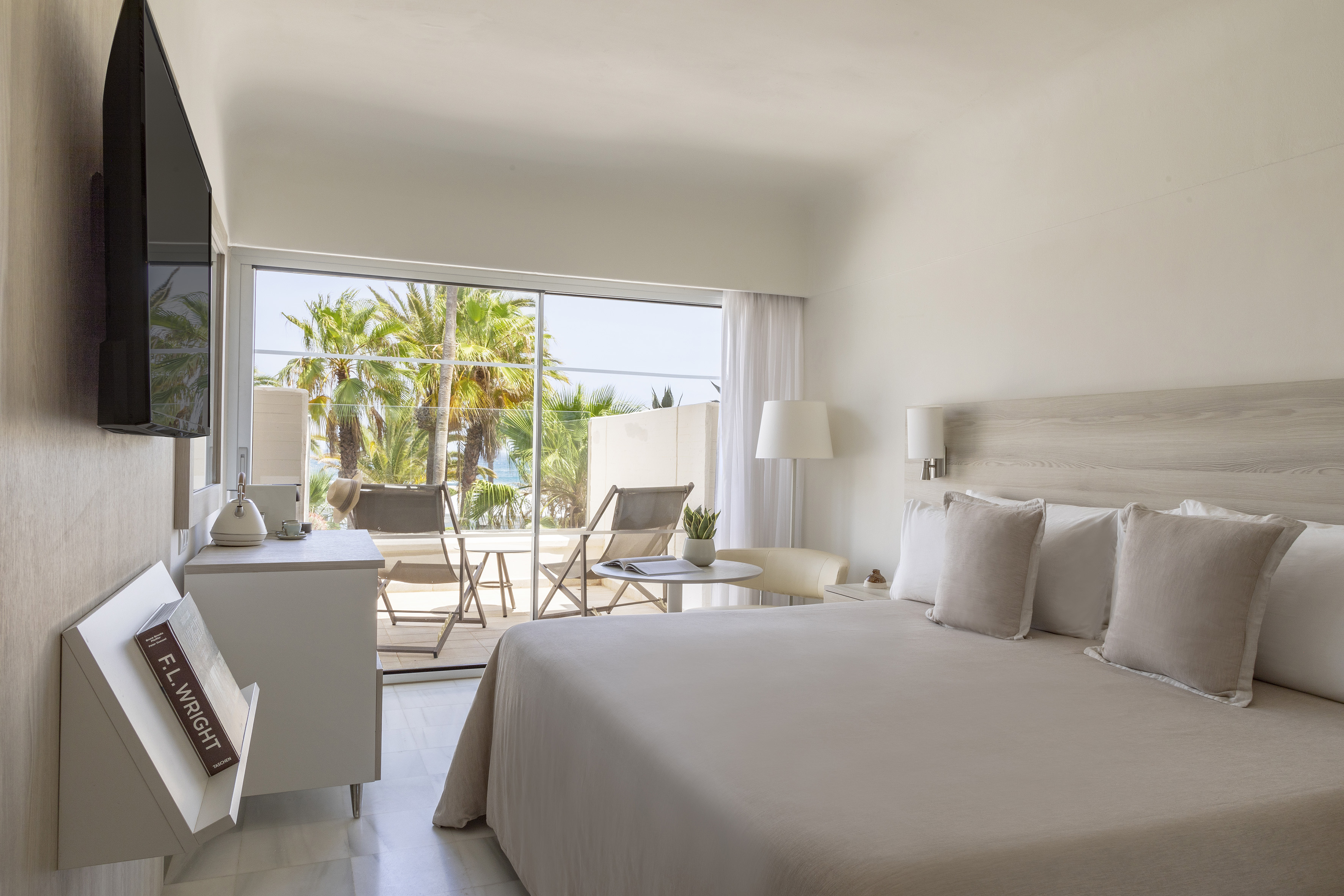 Minimalist hotel bedroom with balcony, ocean view, and tropical palm trees.