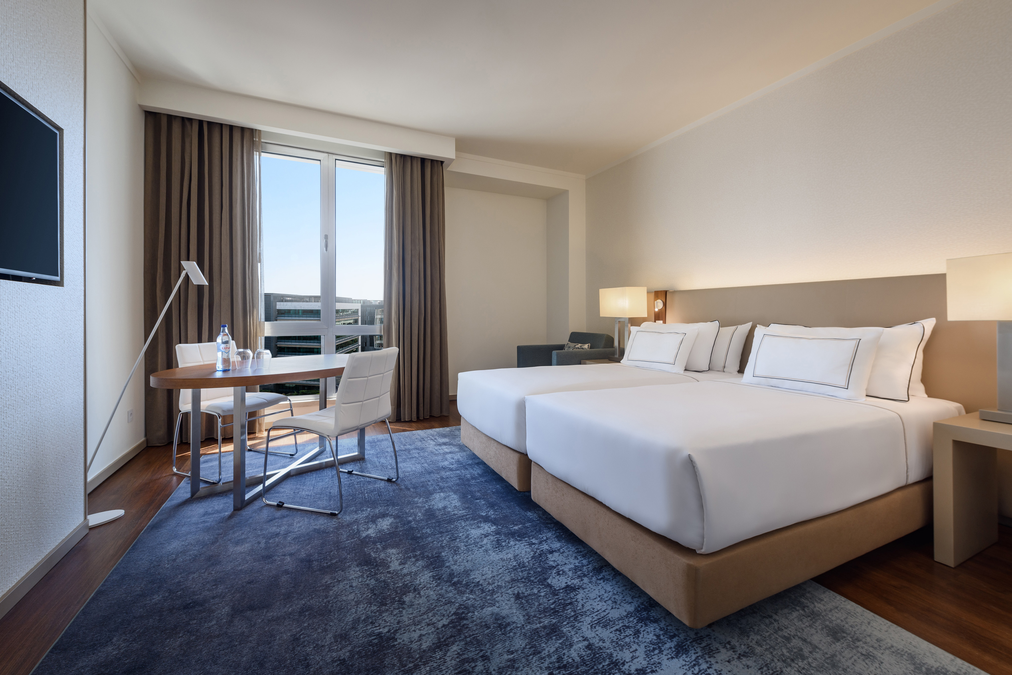 Hotel room with twin beds, blue rug, desk, and large window.