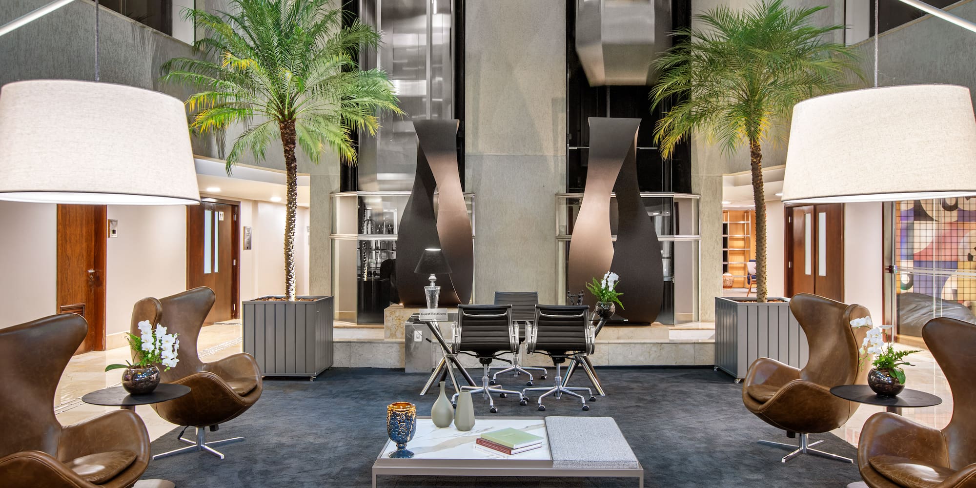 Modern hotel lobby lounge with brown leather chairs, palm trees, and elevators.