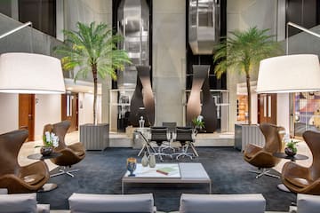 Modern hotel lobby lounge with brown leather chairs, palm trees, and elevators.