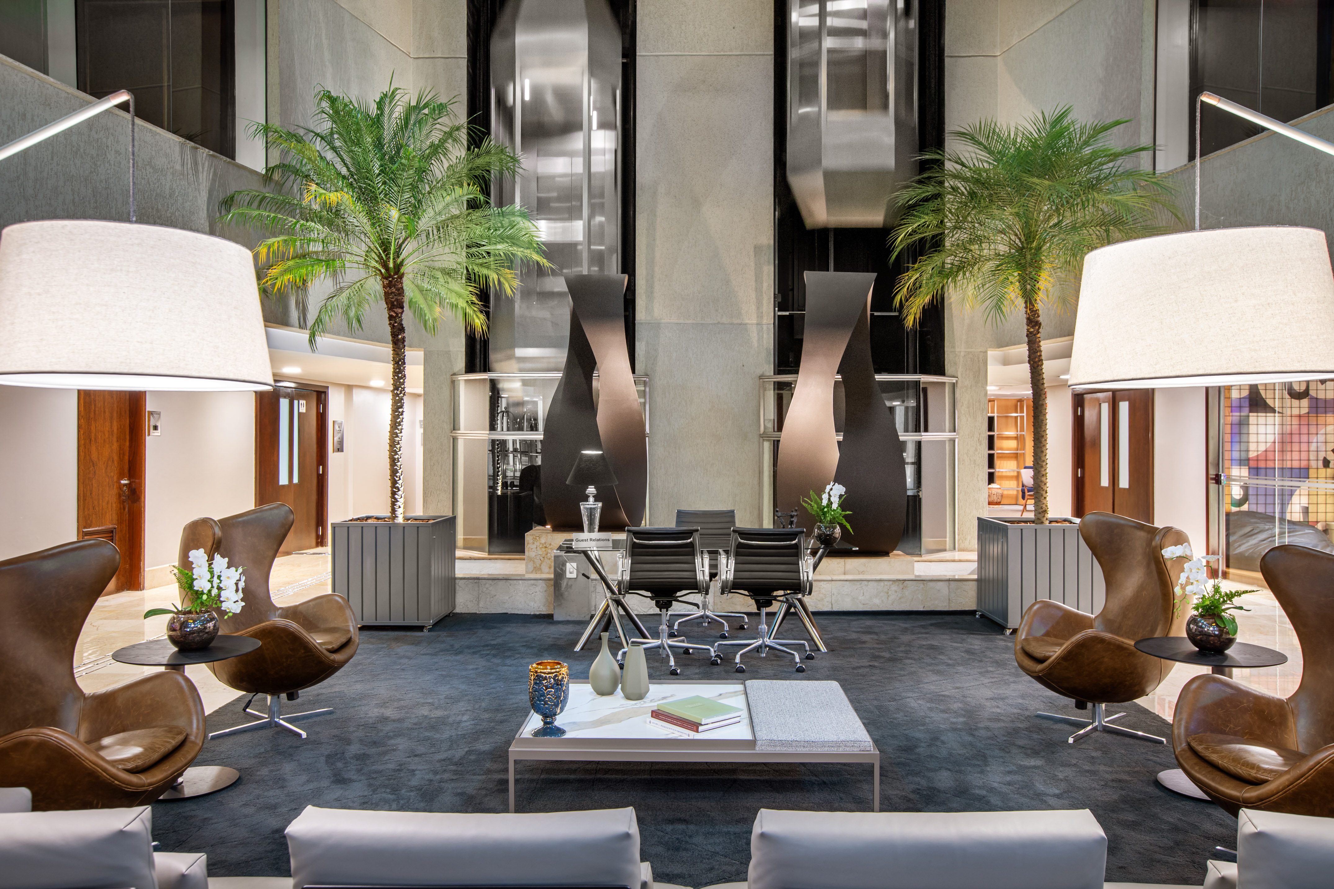 Modern hotel lobby lounge with brown leather chairs, palm trees, and elevators.