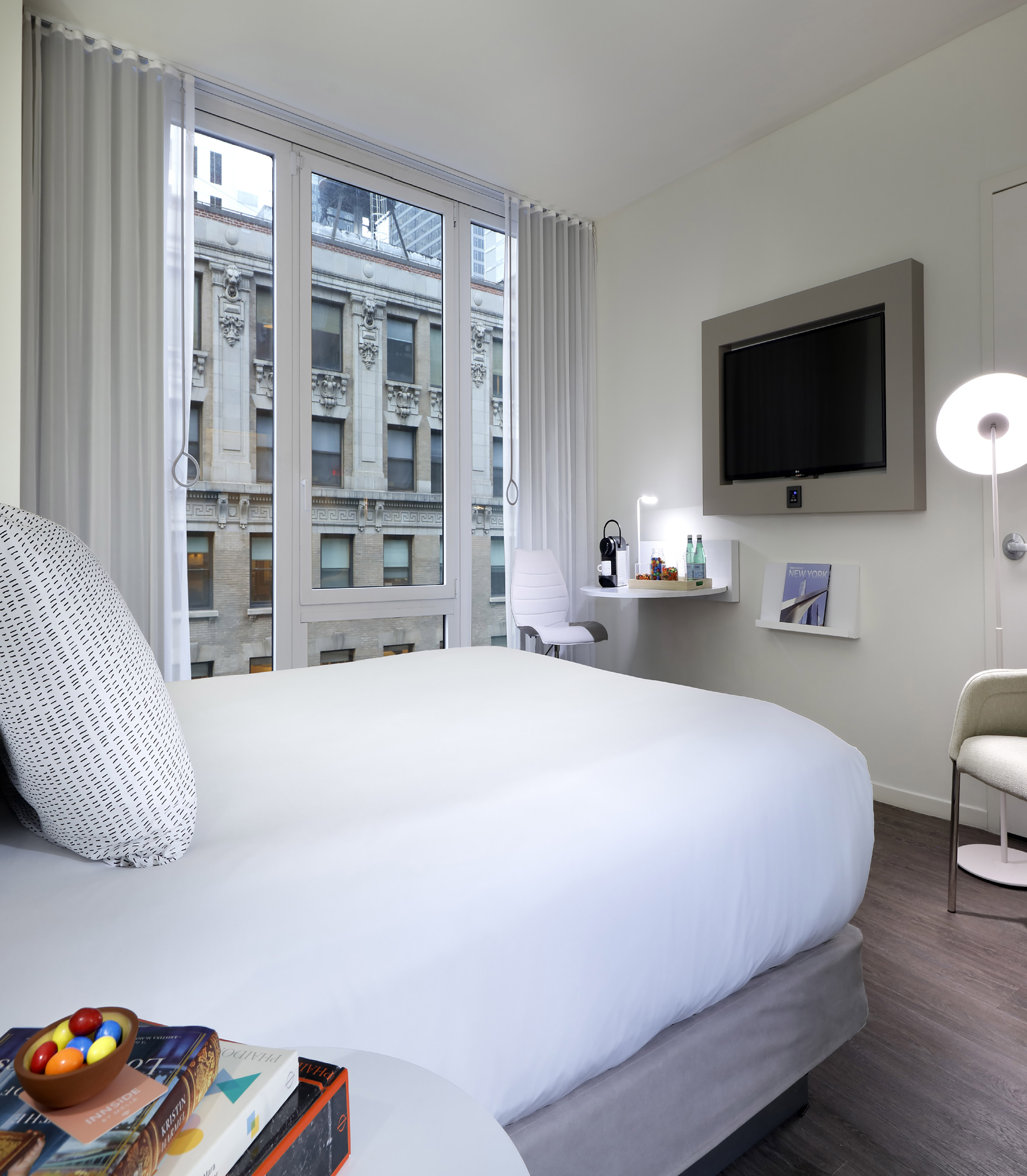 Modern hotel bedroom with city view window, white bed, and wall TV.