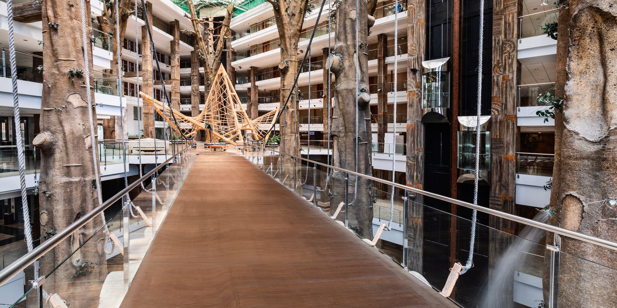 Hotel atrium with wooden bridge, tree-like columns, and glass elevator.