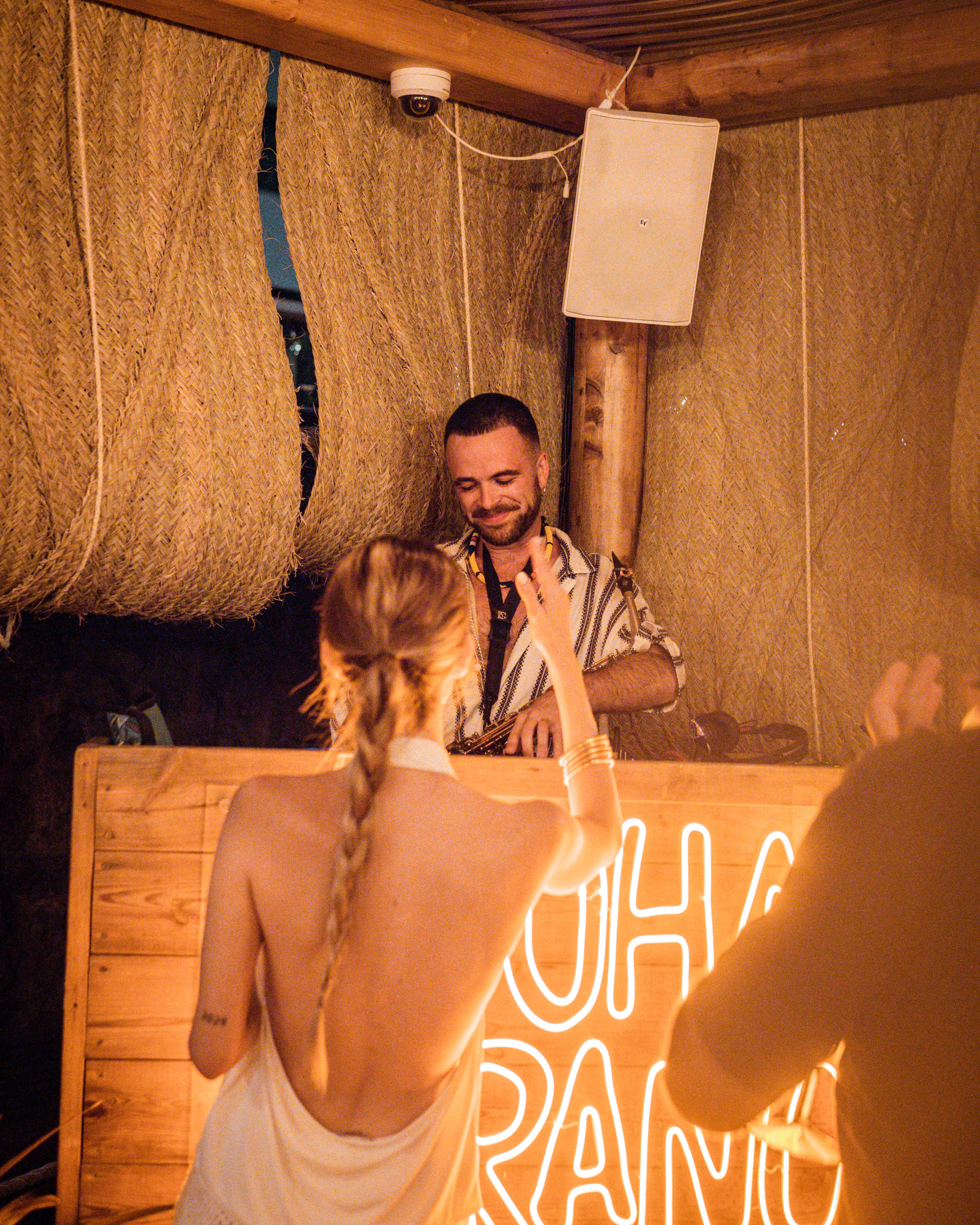 Man DJing, woman dancing at a tropical bar with a neon sign.