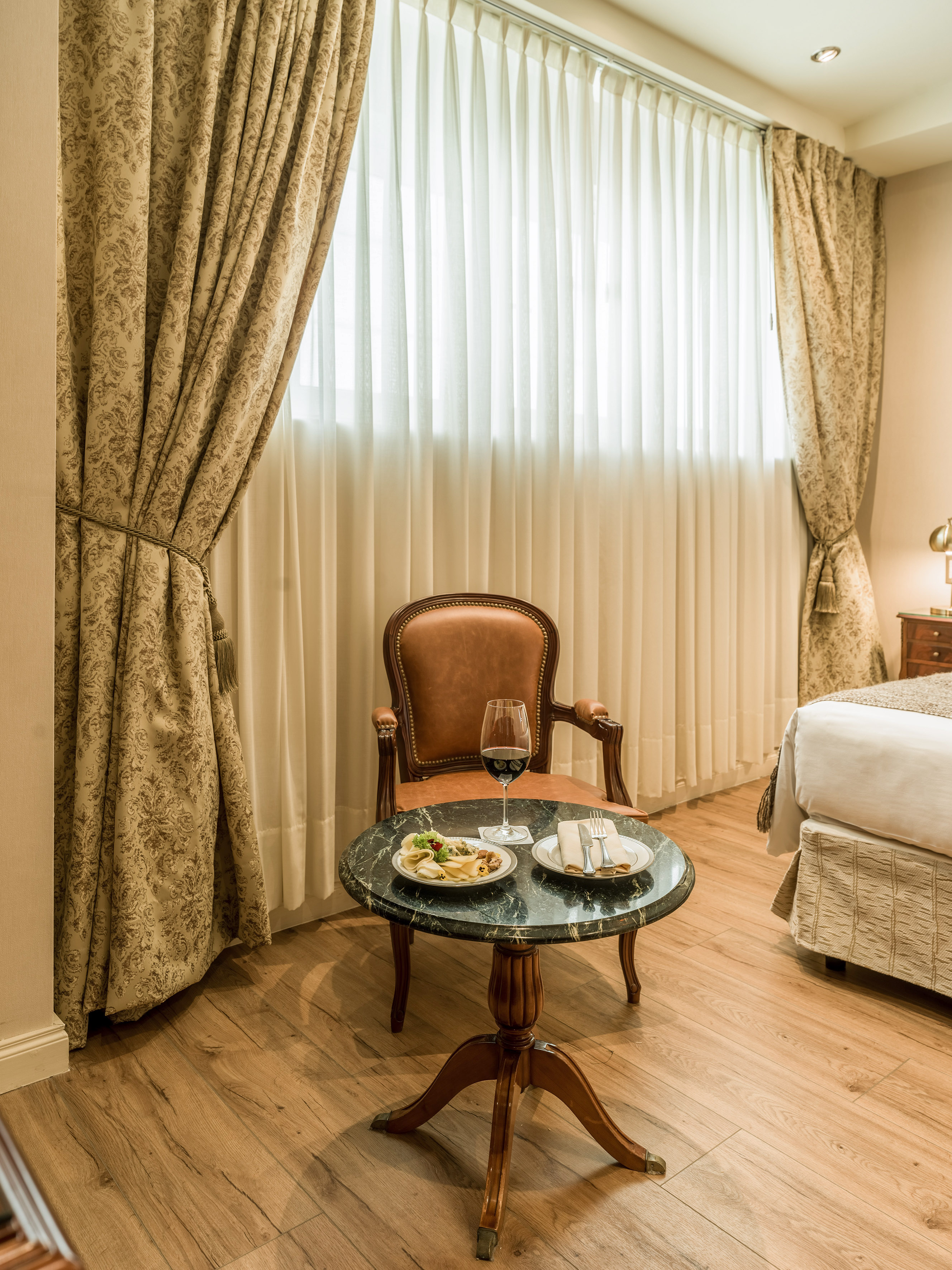 Hotel room: armchair, marble table with wine and snacks, patterned curtains.