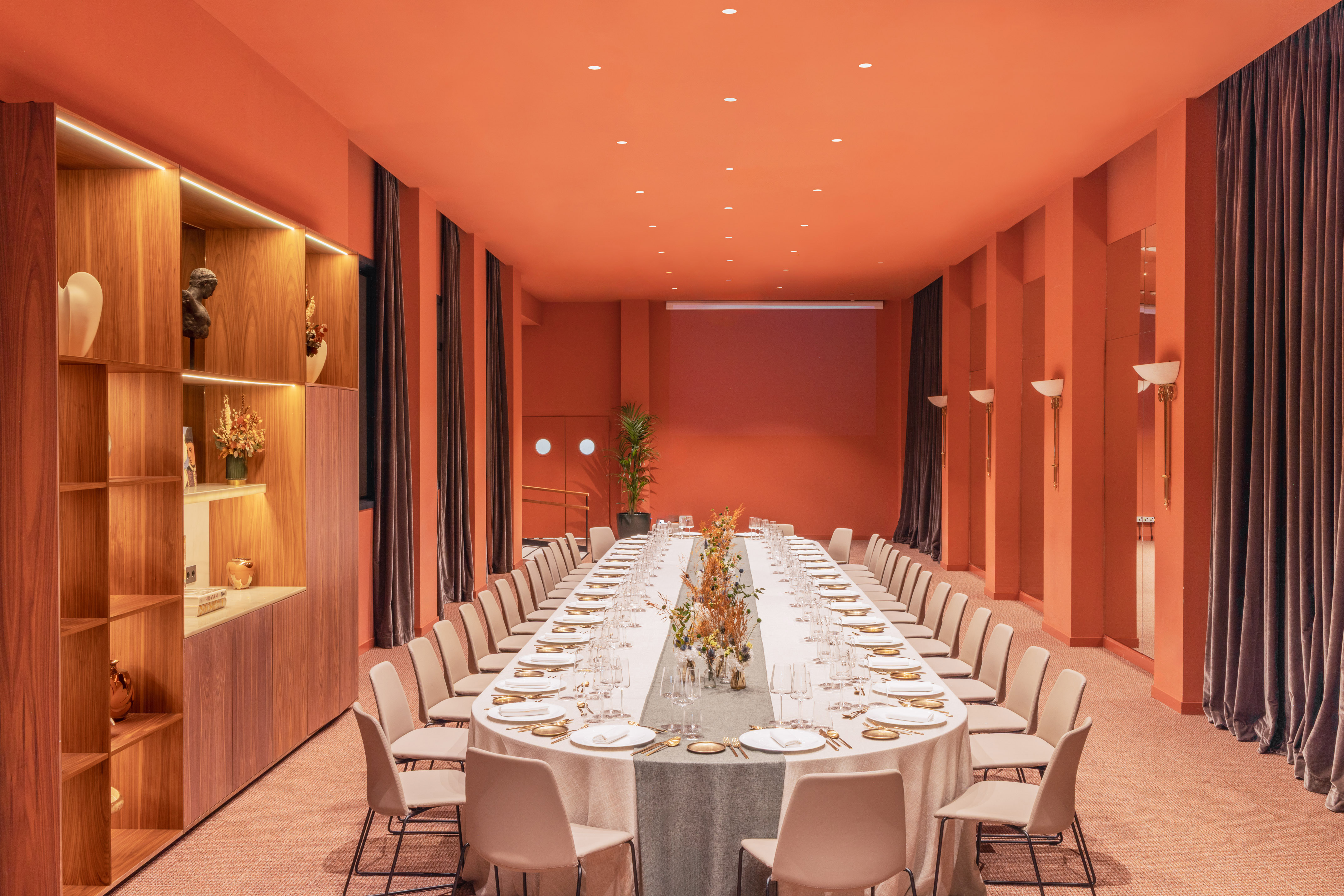 Elegant private dining room with long oval table, orange walls, and wooden shelving.
