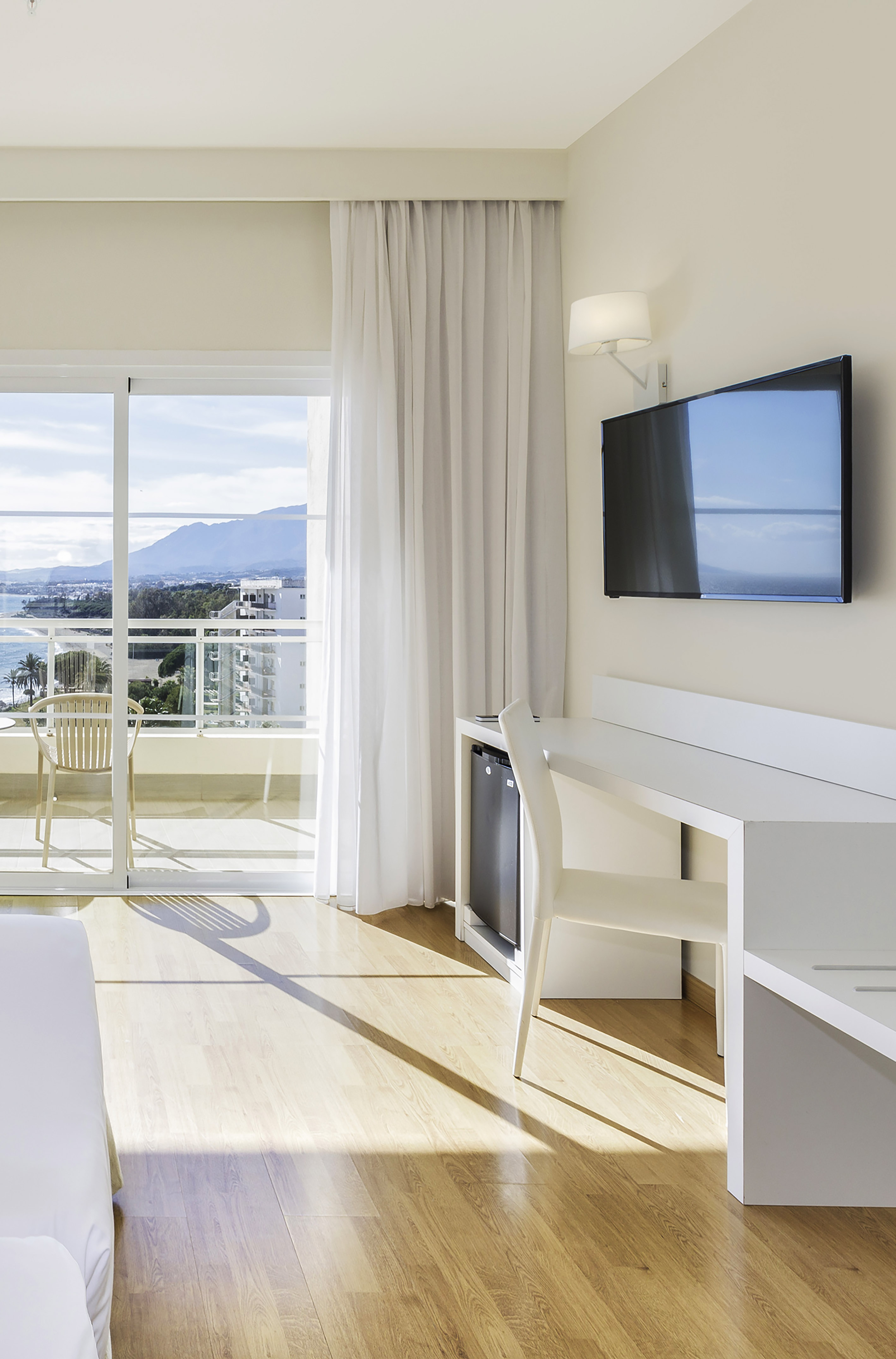 Modern hotel room with balcony, sea and mountain view, desk, and TV.
