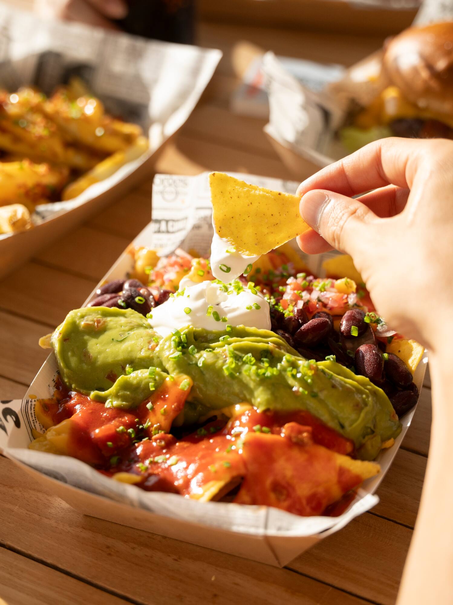 Hand dipping chip into loaded nachos with guacamole and sour cream, outdoor fast food.
