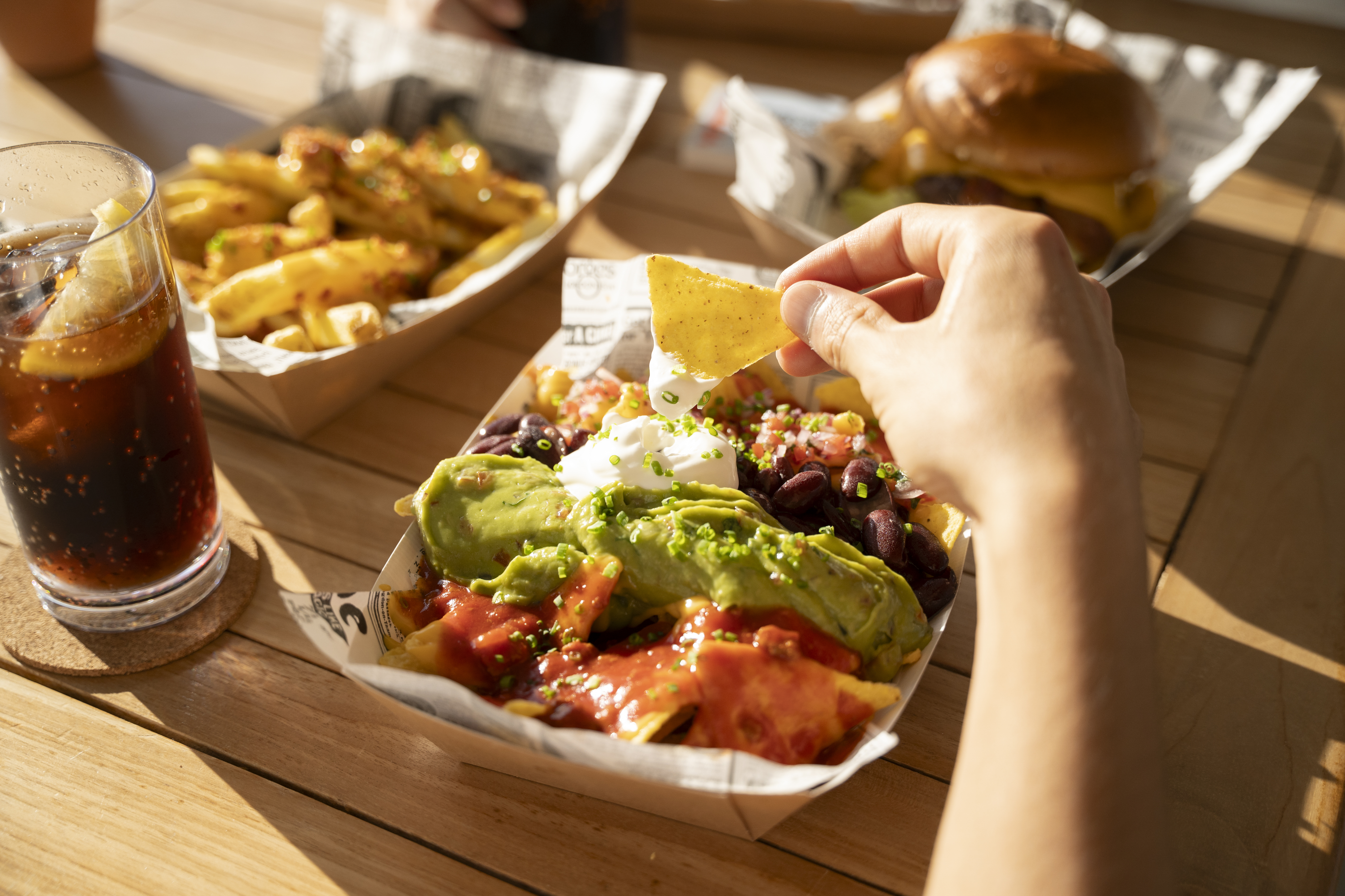Hand dipping chip into loaded nachos with guacamole and sour cream, outdoor fast food.